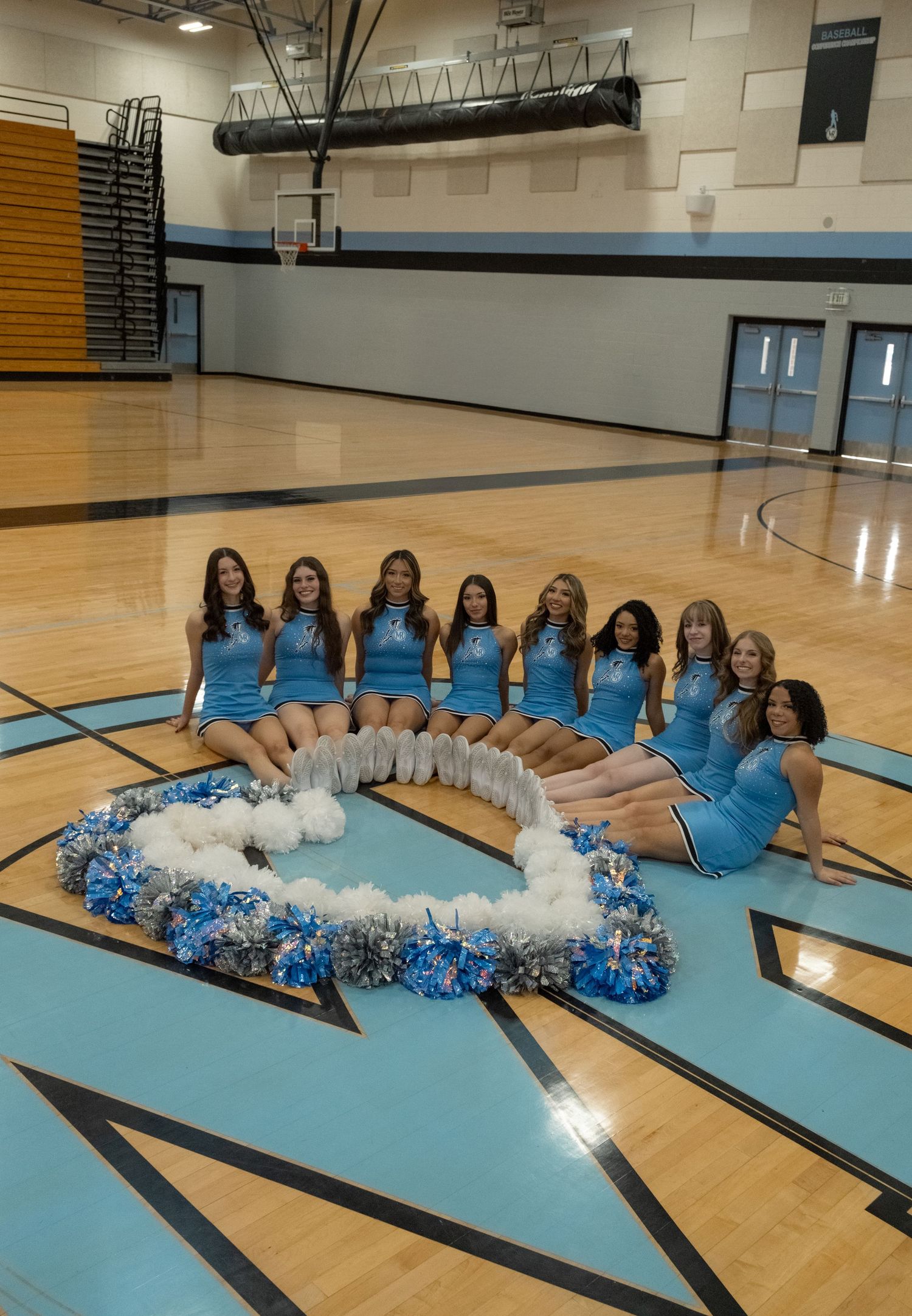 High school cheerleaders in blue uniforms pose with pom-poms on a gymnasium basketball court floor.