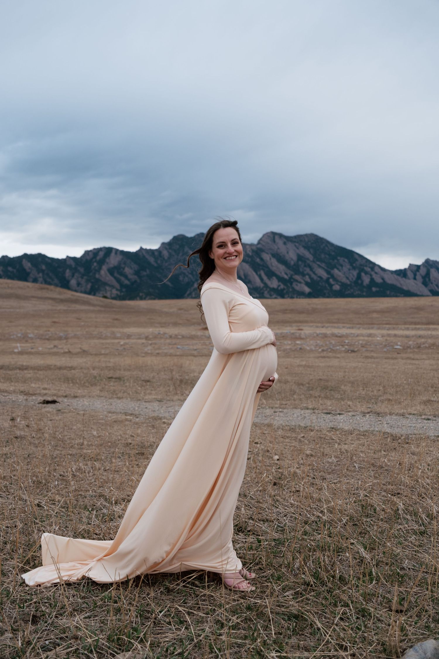 Pregnant woman in elegant peach gown poses in Colorado field with Flatirons mountains backdrop.