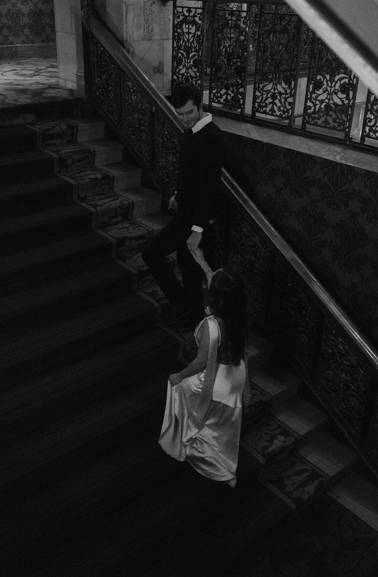 Black and white photo of a couple on ornate stairs, woman in elegant gown, man in dark suit, viewed from above.