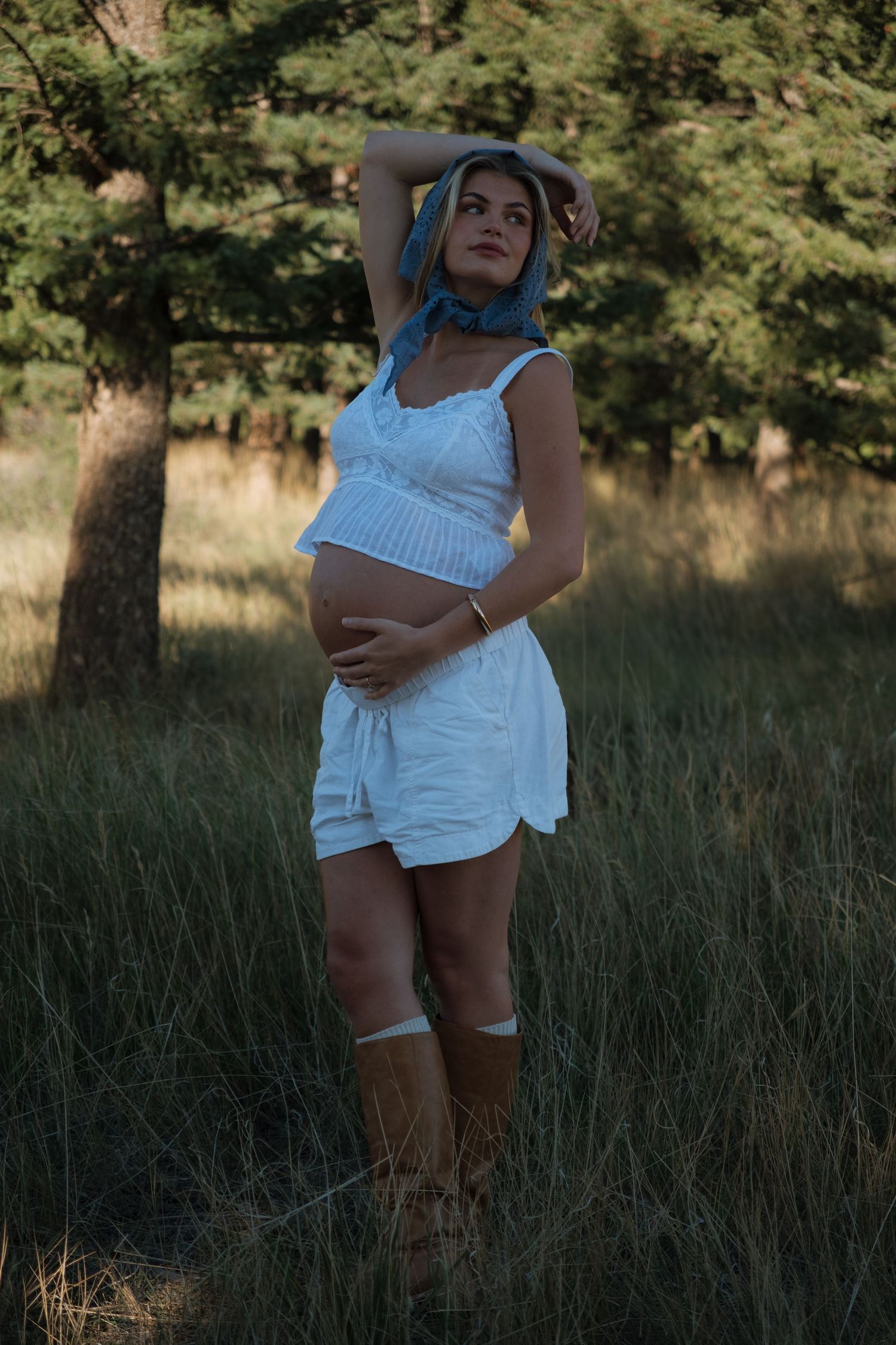 Pregnant woman in a white crop top, shorts, blue headscarf and brown boots posing in a grassy field beside tall pine trees.