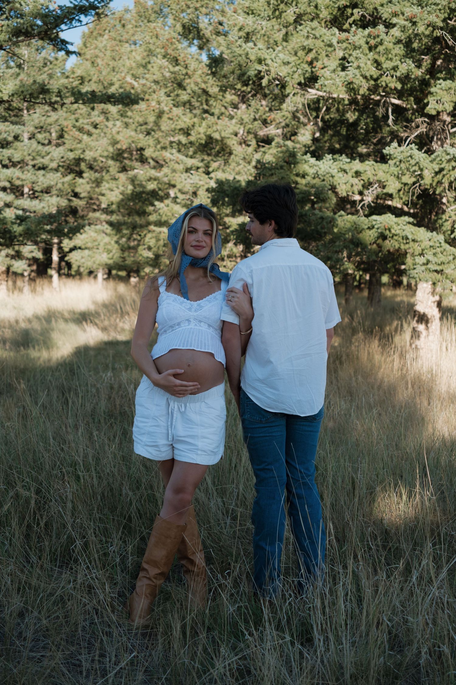 Pregnant woman in white top, shorts, and blue headscarf holds her belly beside a man in a white shirt and jeans in a sunny grassy field.