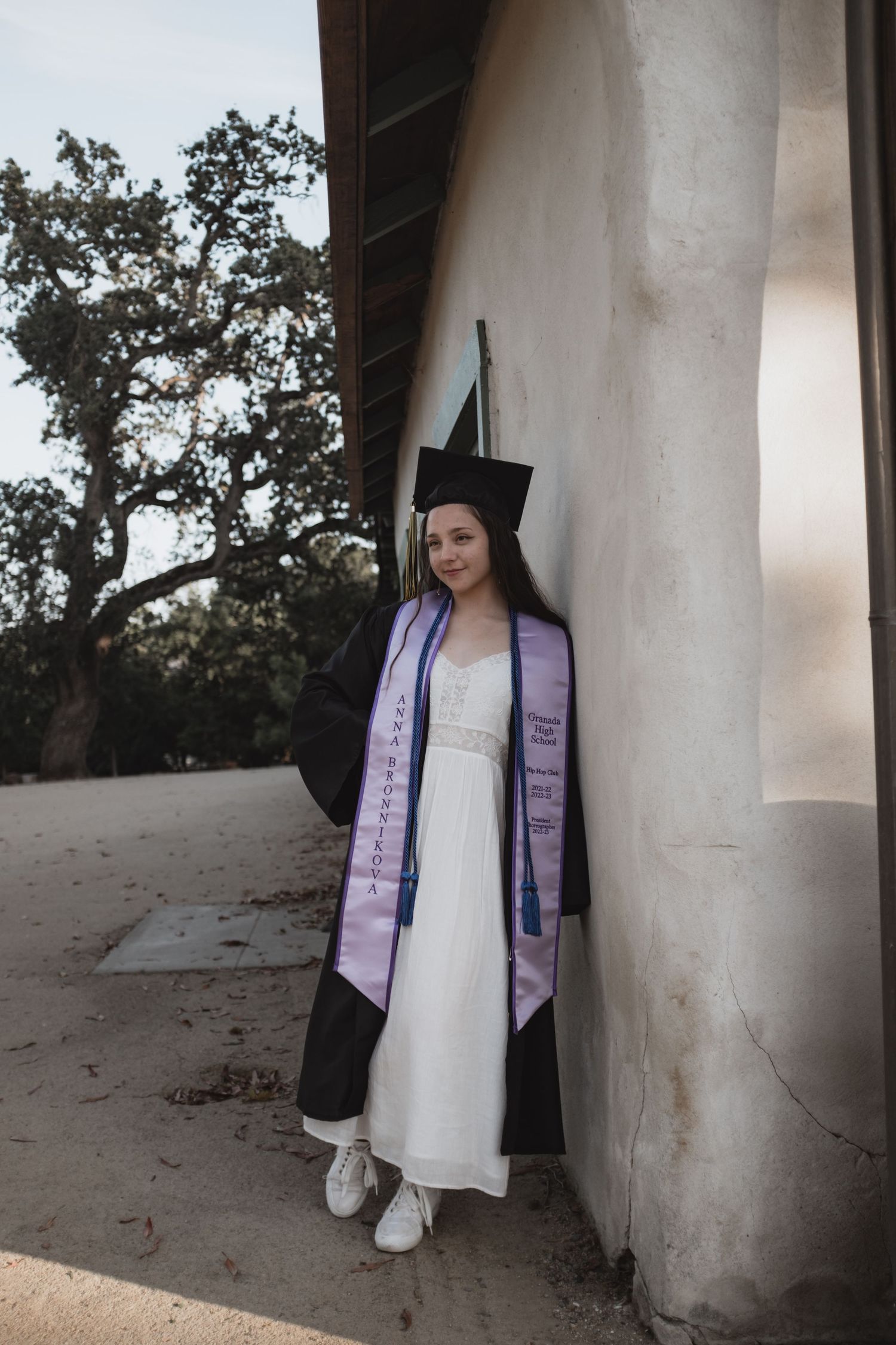 Graduate in cap and gown poses against a white column wearing purple graduation stoles.