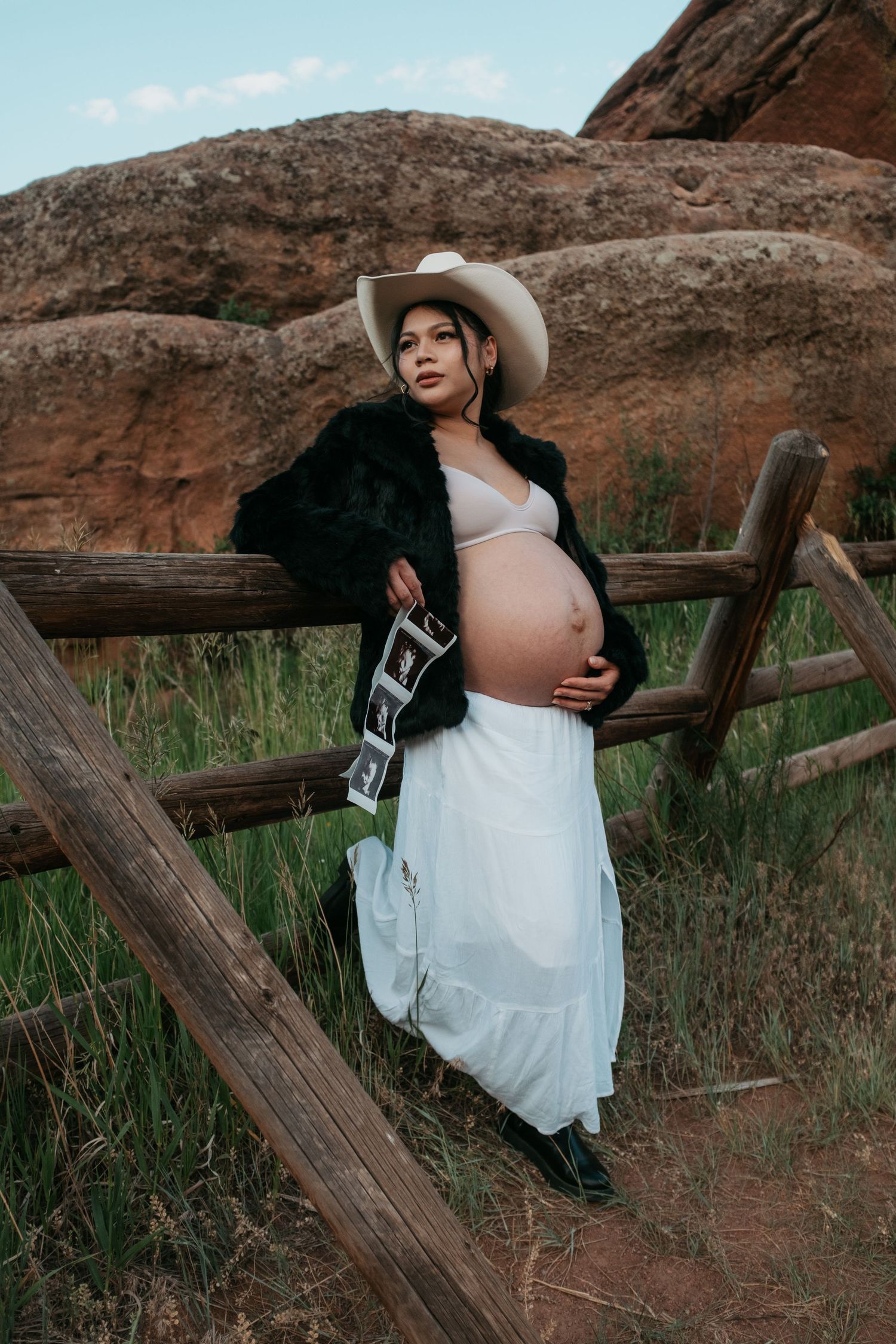 Pregnant woman in cowboy hat poses with ultrasound photos by wooden fence.