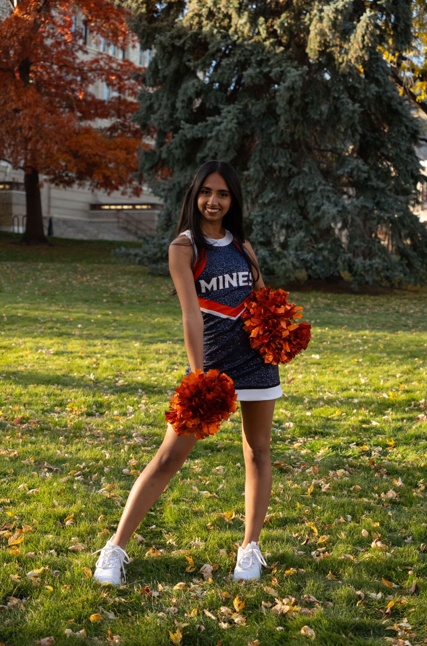 Young woman in Mines cheerleader uniform holding orange pom-poms on a grass field with autumn trees.