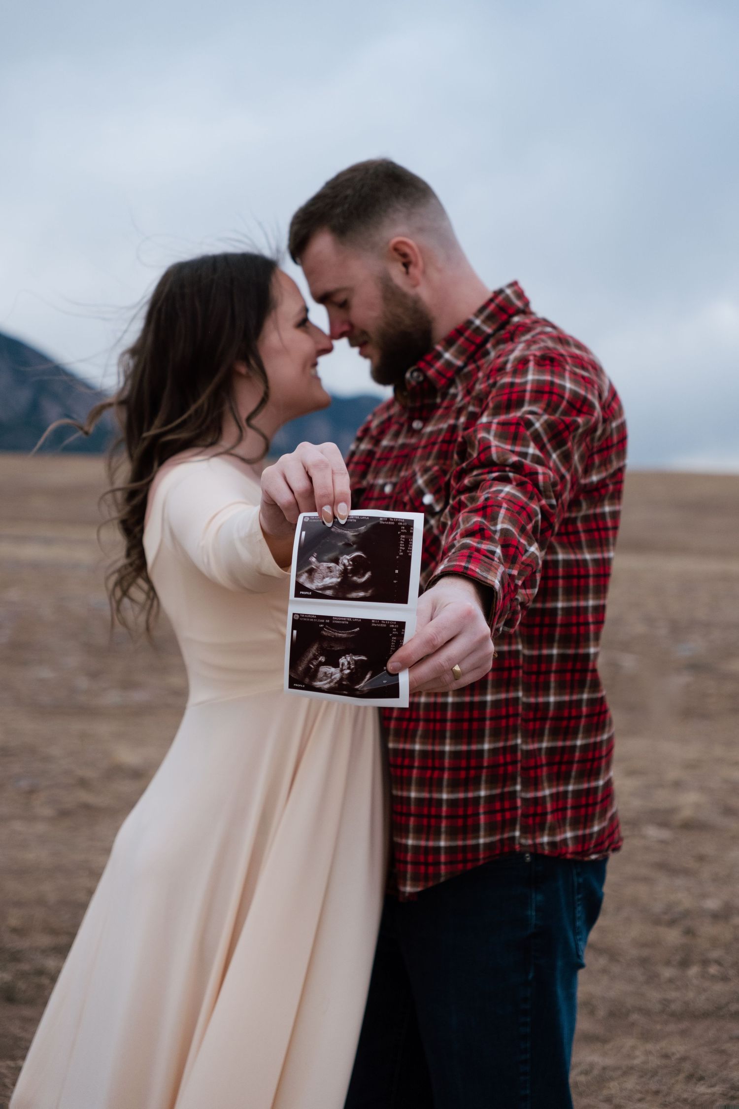 Couple holding ultrasound photos outdoors in a field, touching foreheads in a tender pregnancy announcement moment.