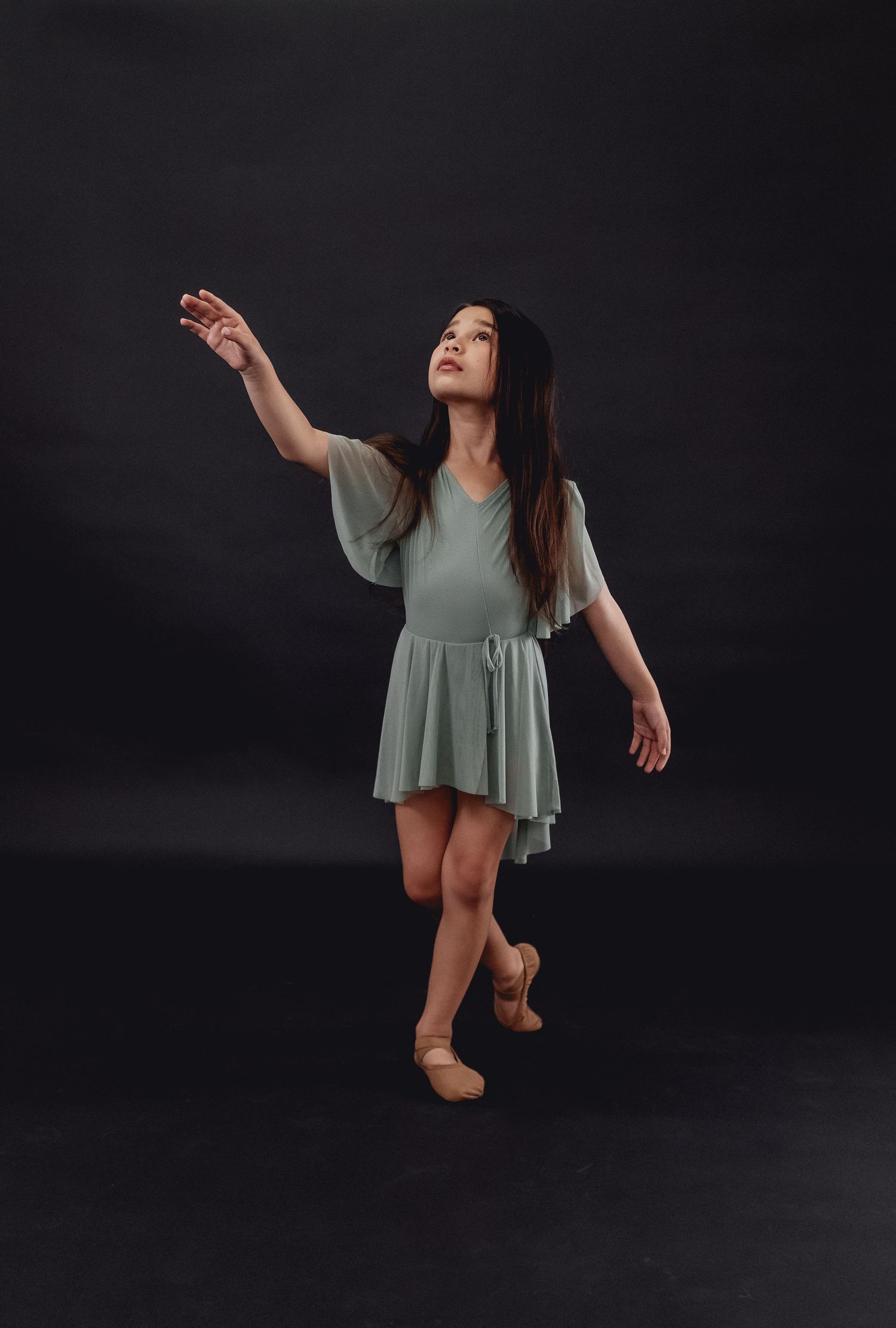 Dancer in flowy sage dress performs graceful movement on dark stage.