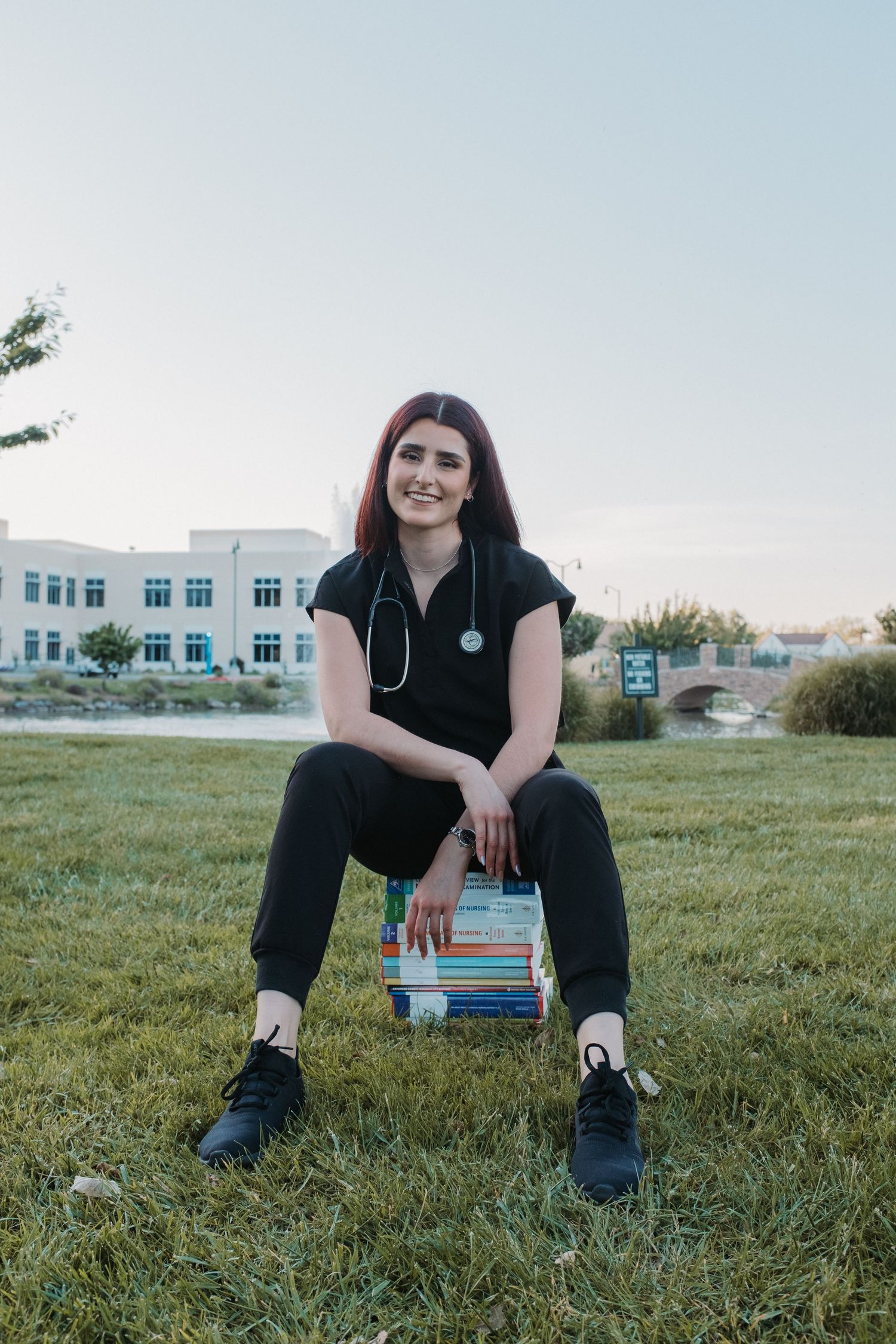 Medical student in scrubs sitting on textbooks outdoors on campus.