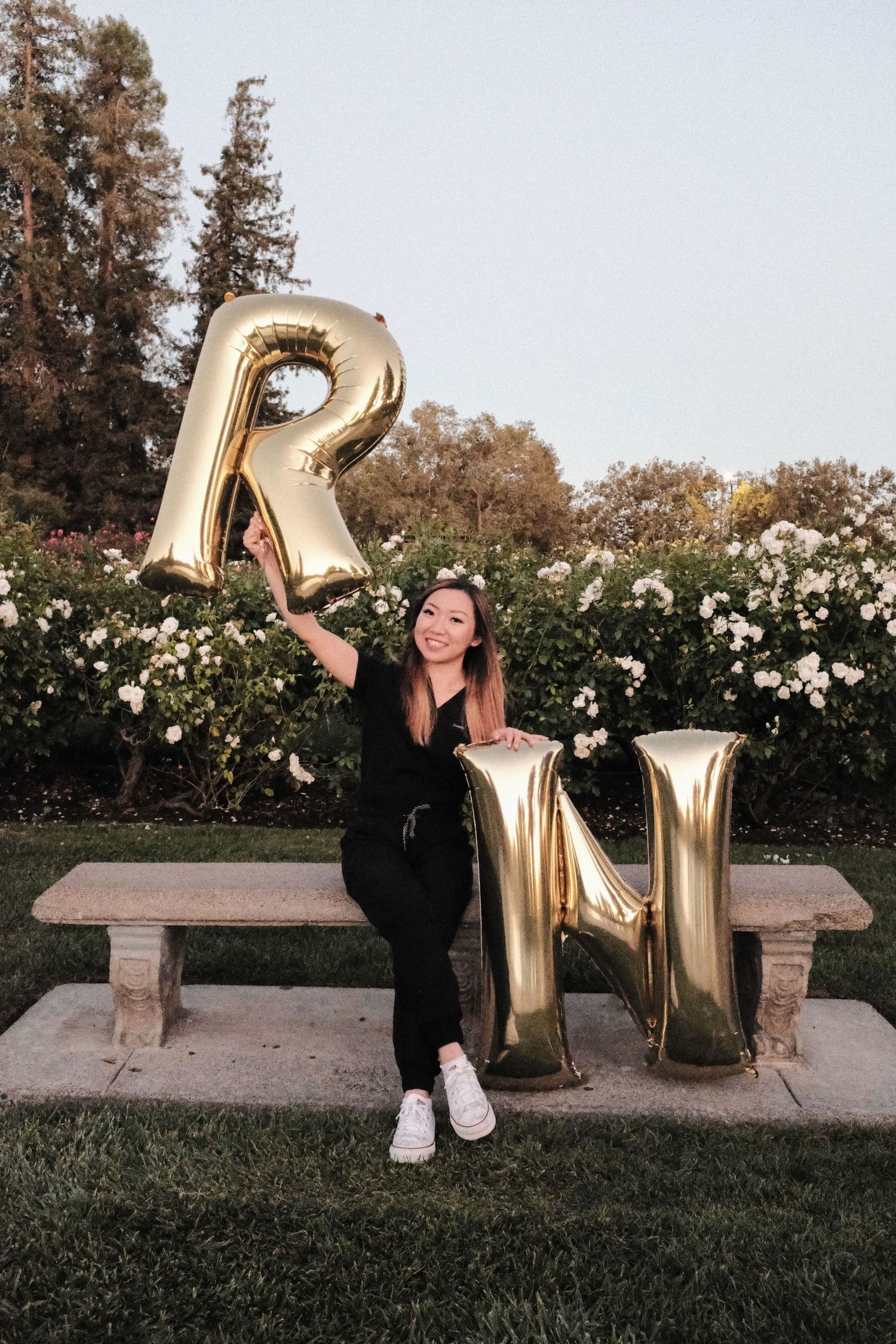 Graduate in black attire posing with large gold RN balloon letters in a garden setting.