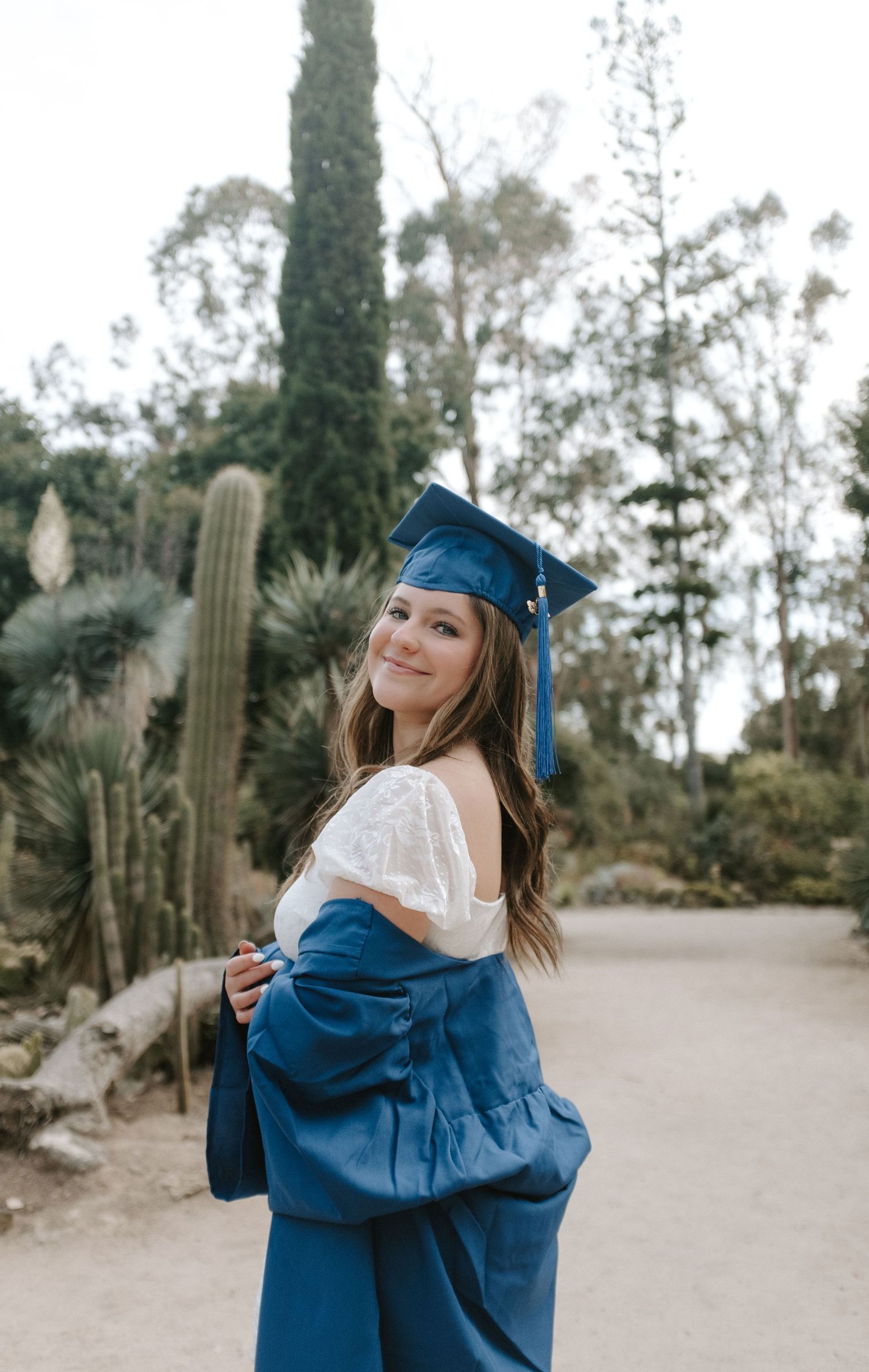 A graduate in blue cap and gown poses in a garden with cactus plants.
