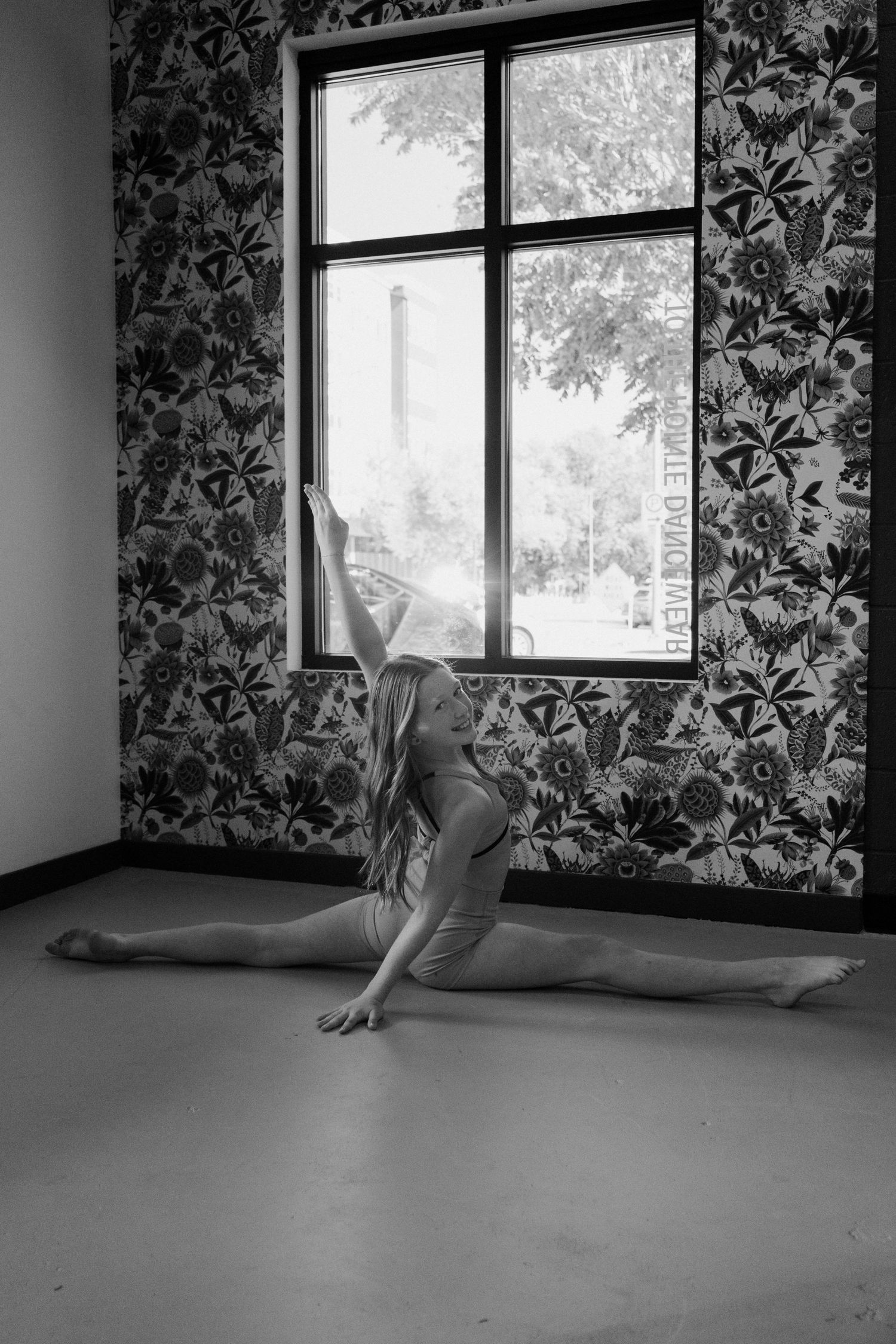Black and white photo of a flexible dancer performing a split on studio floor before floral wallpaper and window.