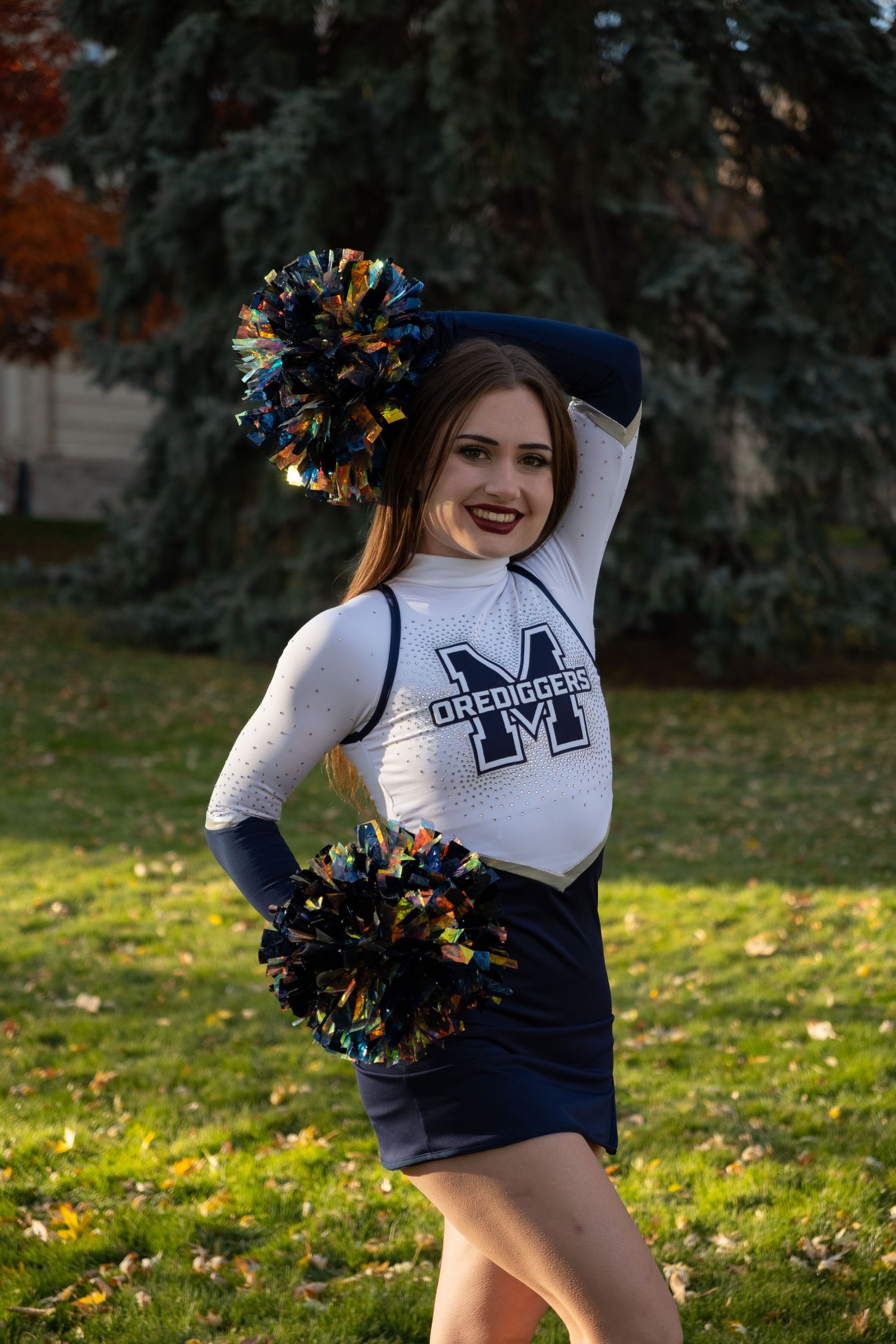 A cheerleader in a white and navy Orediggers uniform poses with colorful pom-poms outdoors on a grass field.