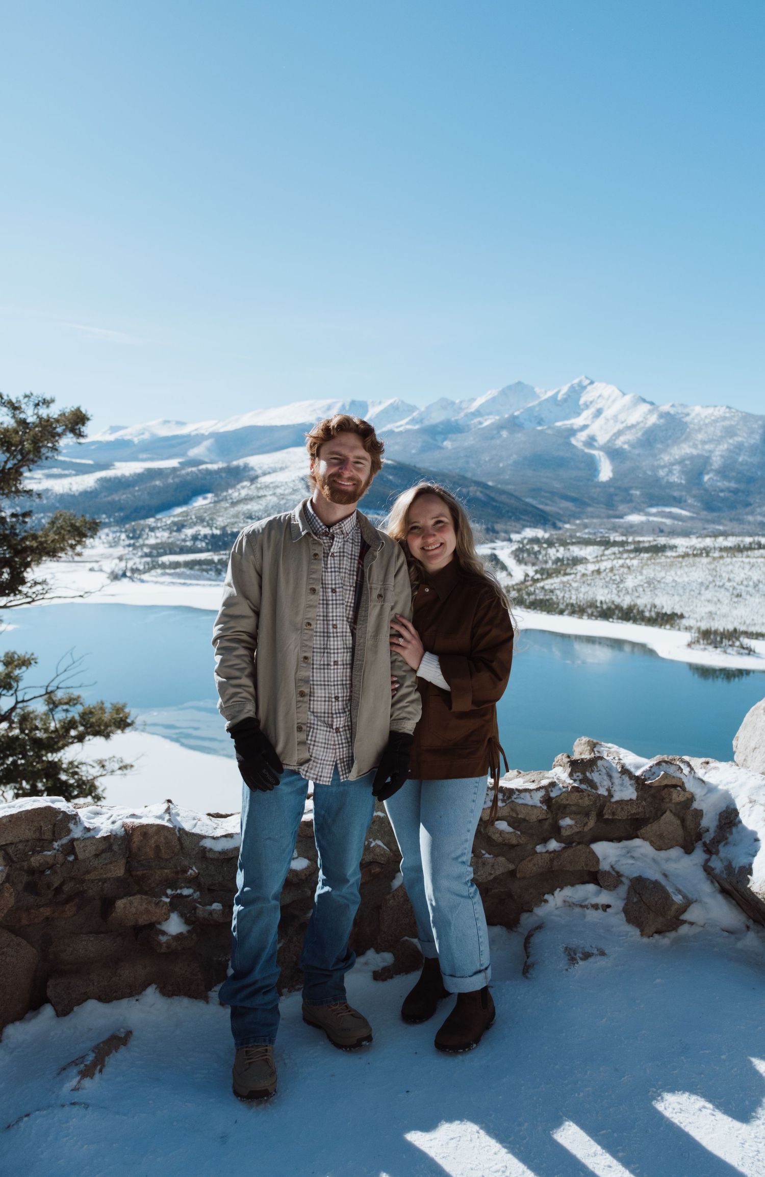 Couple smiling together on a snowy overlook with a mountain lake and snow-capped peaks in the background.