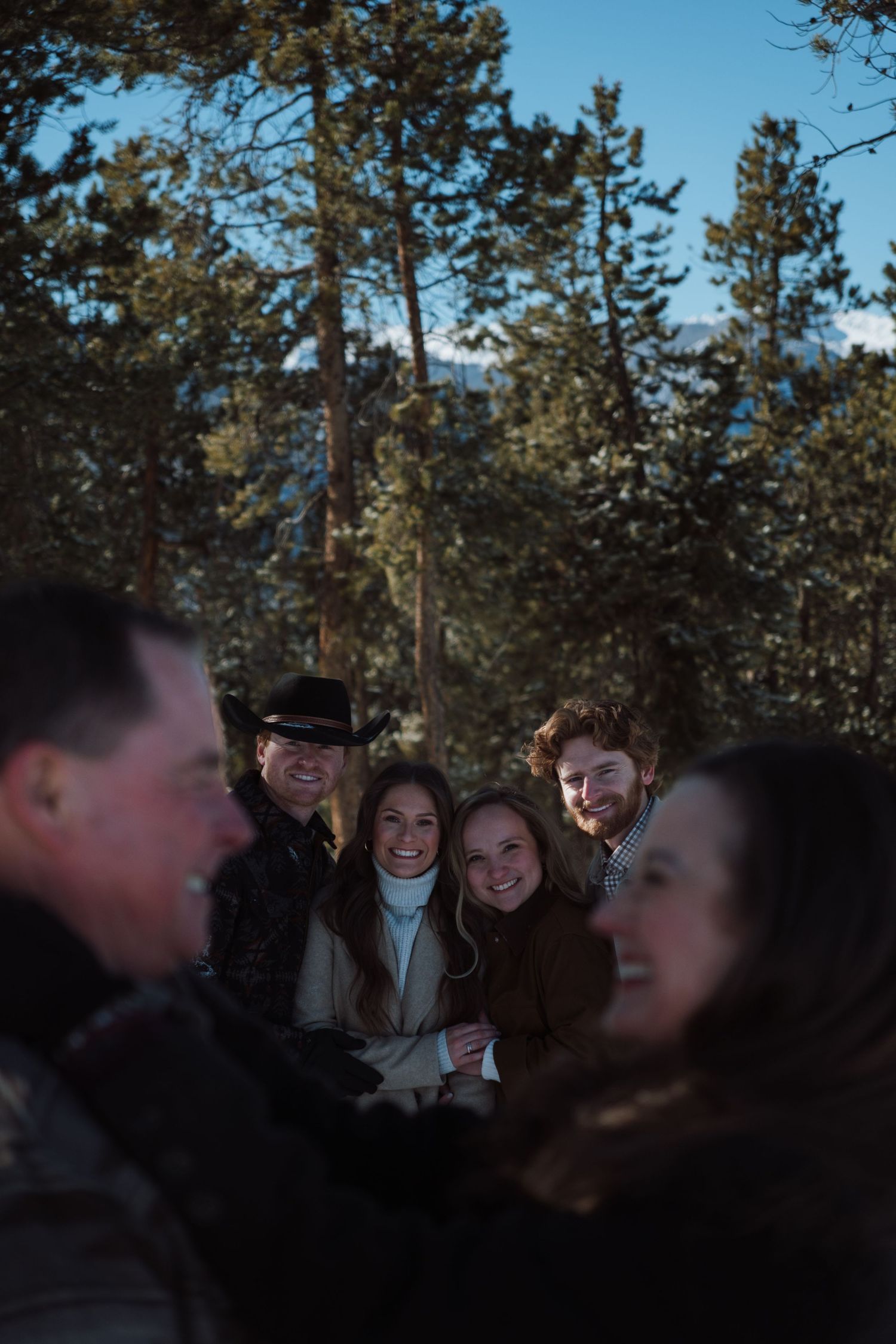 Group of friends smiling outdoors in a winter forest setting with snow-dusted pine trees in the background.
