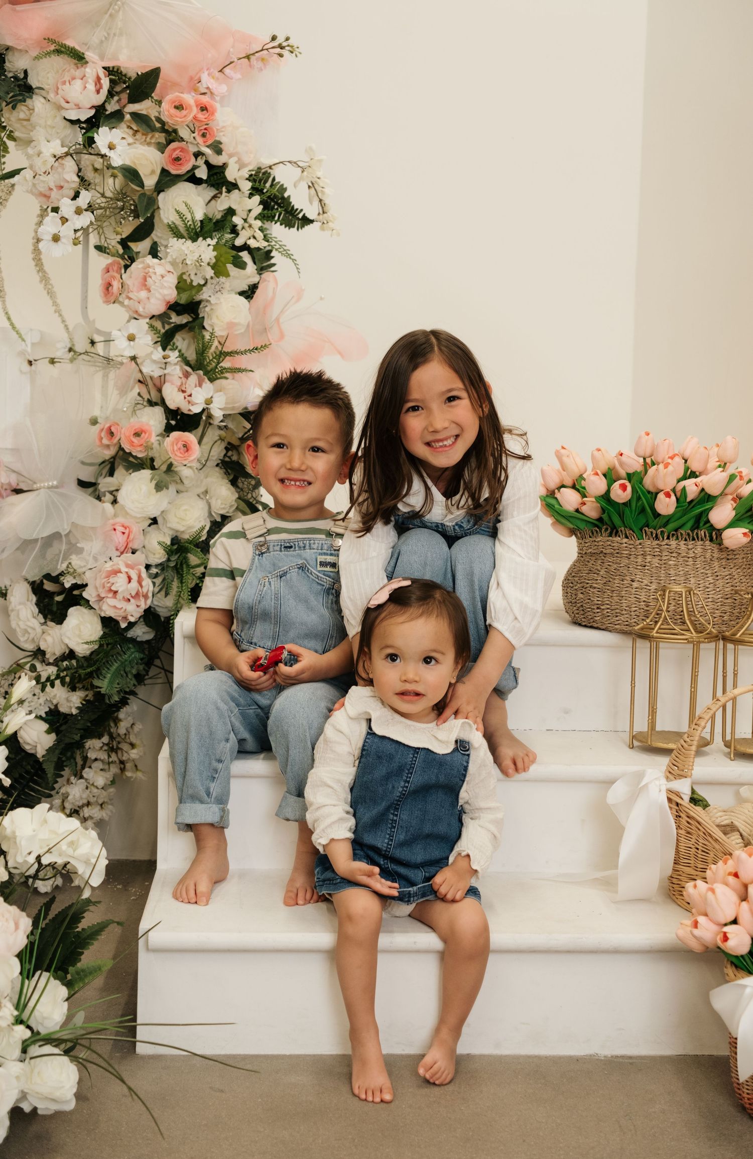 Three young children in denim overalls pose on white steps surrounded by pink and white floral arrangements.