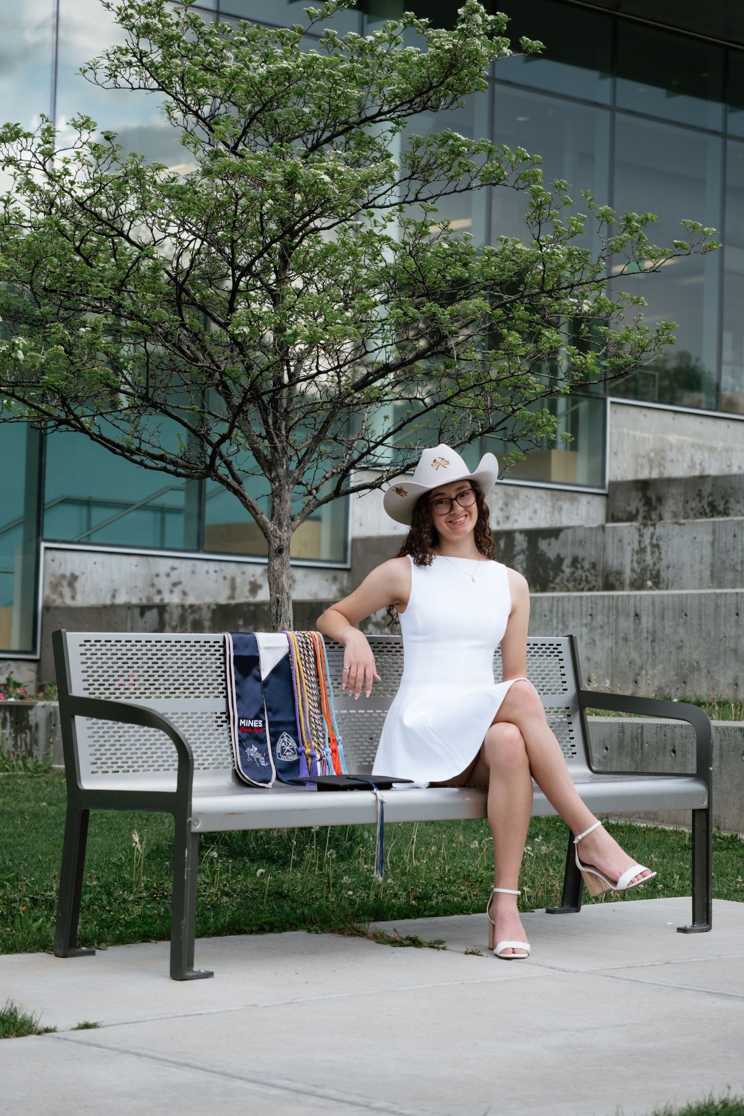 Woman in white dress and cowboy hat sitting on a metal bench outdoors near a modern glass building.