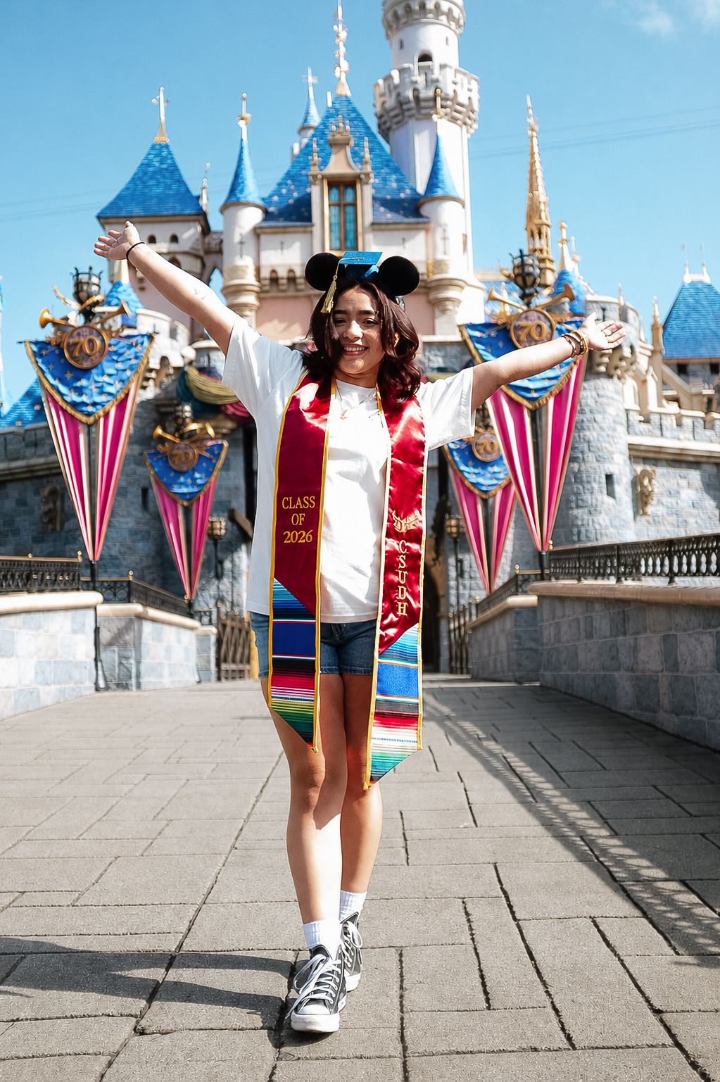 Graduate in Class of 2021 sash and Mickey ears celebrates in front of Disneyland castle with arms raised.