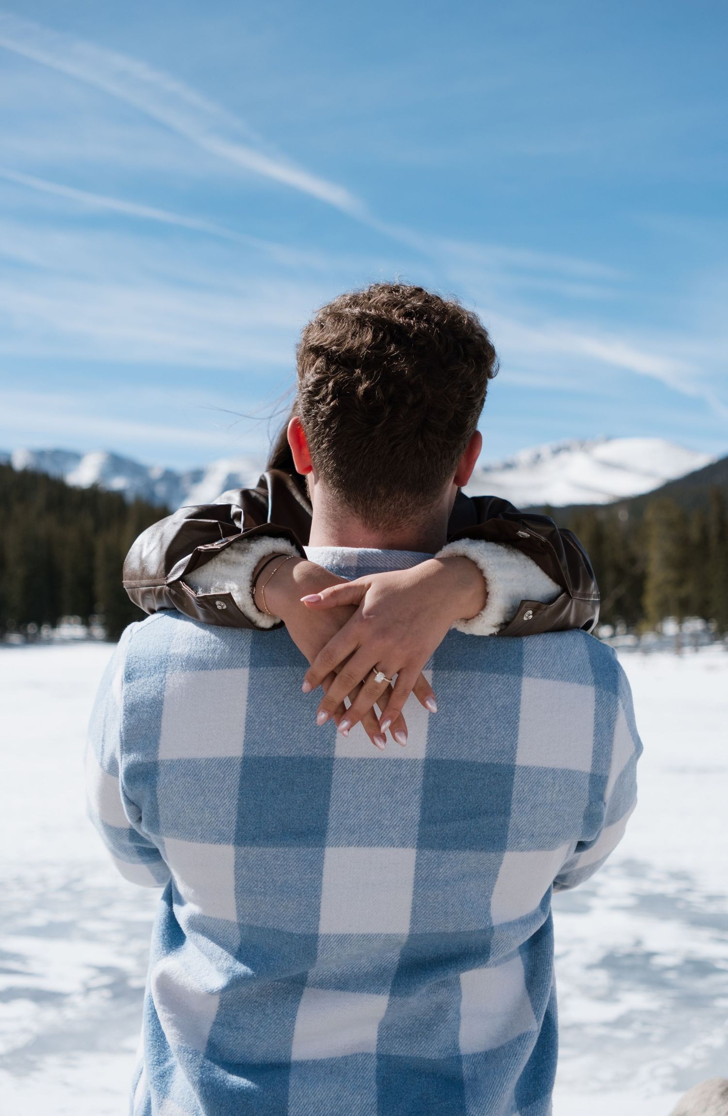 Couple embracing in snowy mountain landscape, woman's hand with engagement ring wrapped around man's neck.