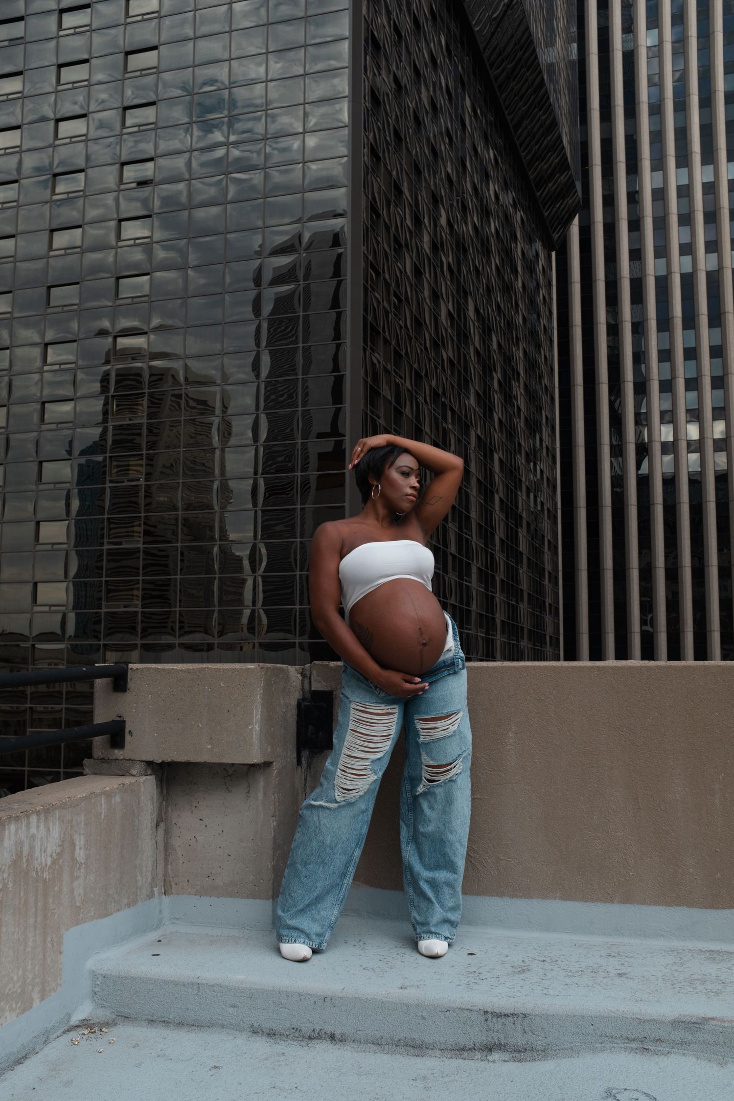 Pregnant woman in white crop top and ripped jeans cradling baby bump on urban steps with glass building backdrop.