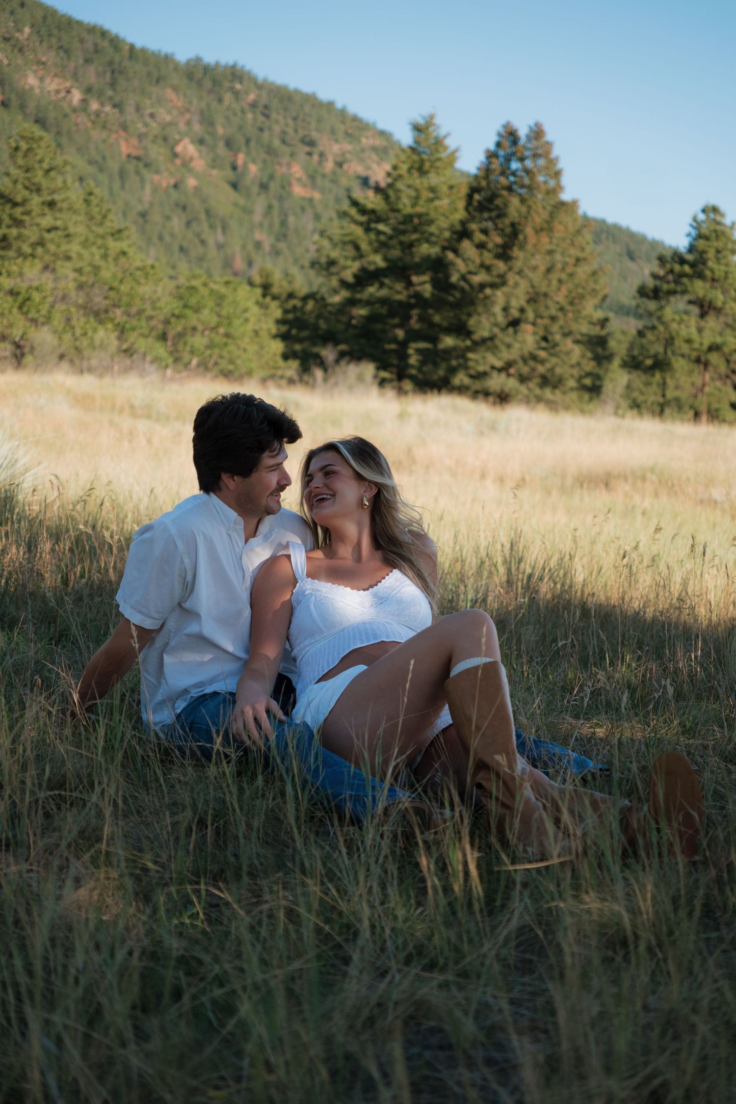 Smiling couple in white outfits sit close together in a sunlit grassy mountain meadow, framed by pine trees and forested hills.