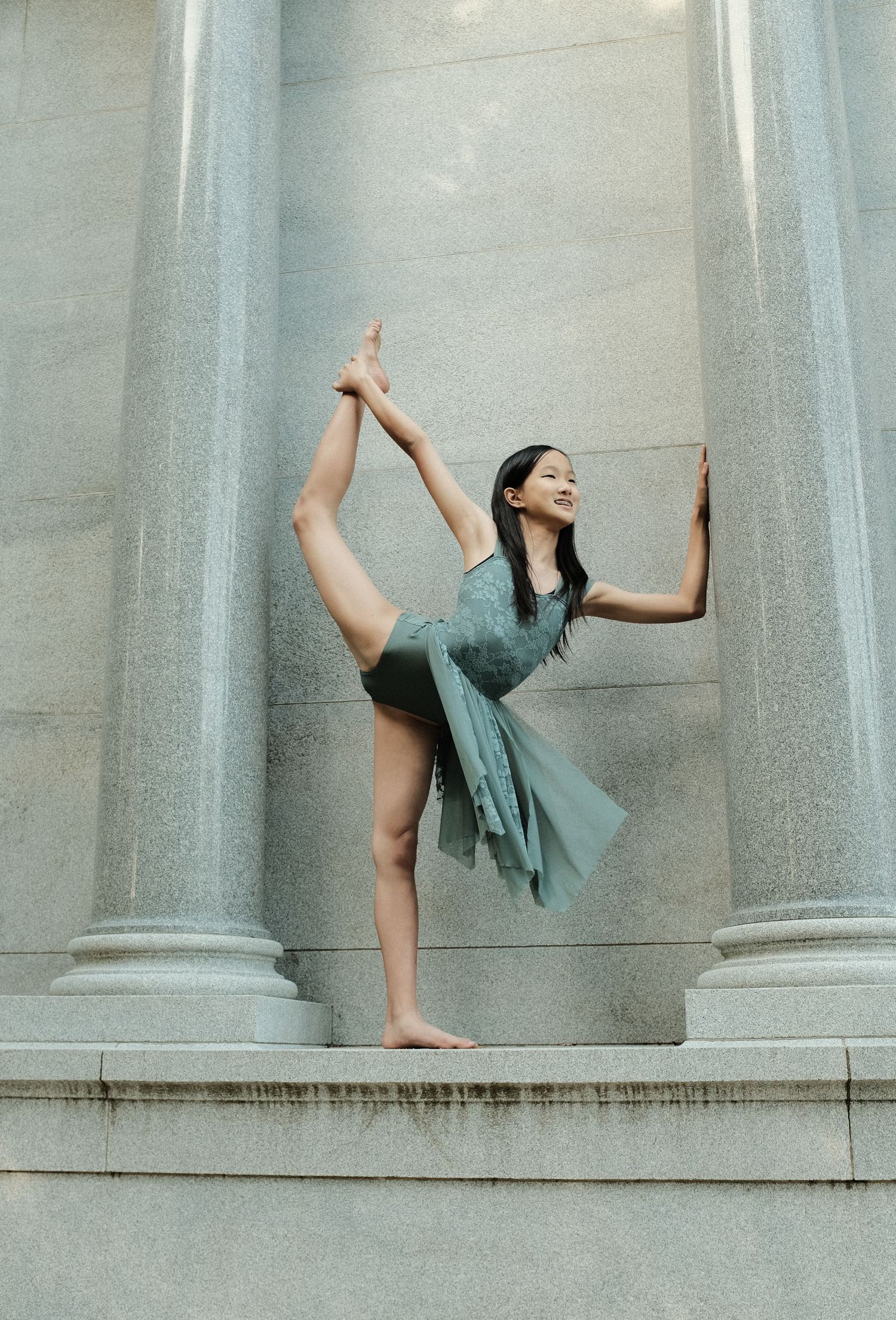 A dancer in a flowy teal dress performs a standing split pose between classical stone columns.