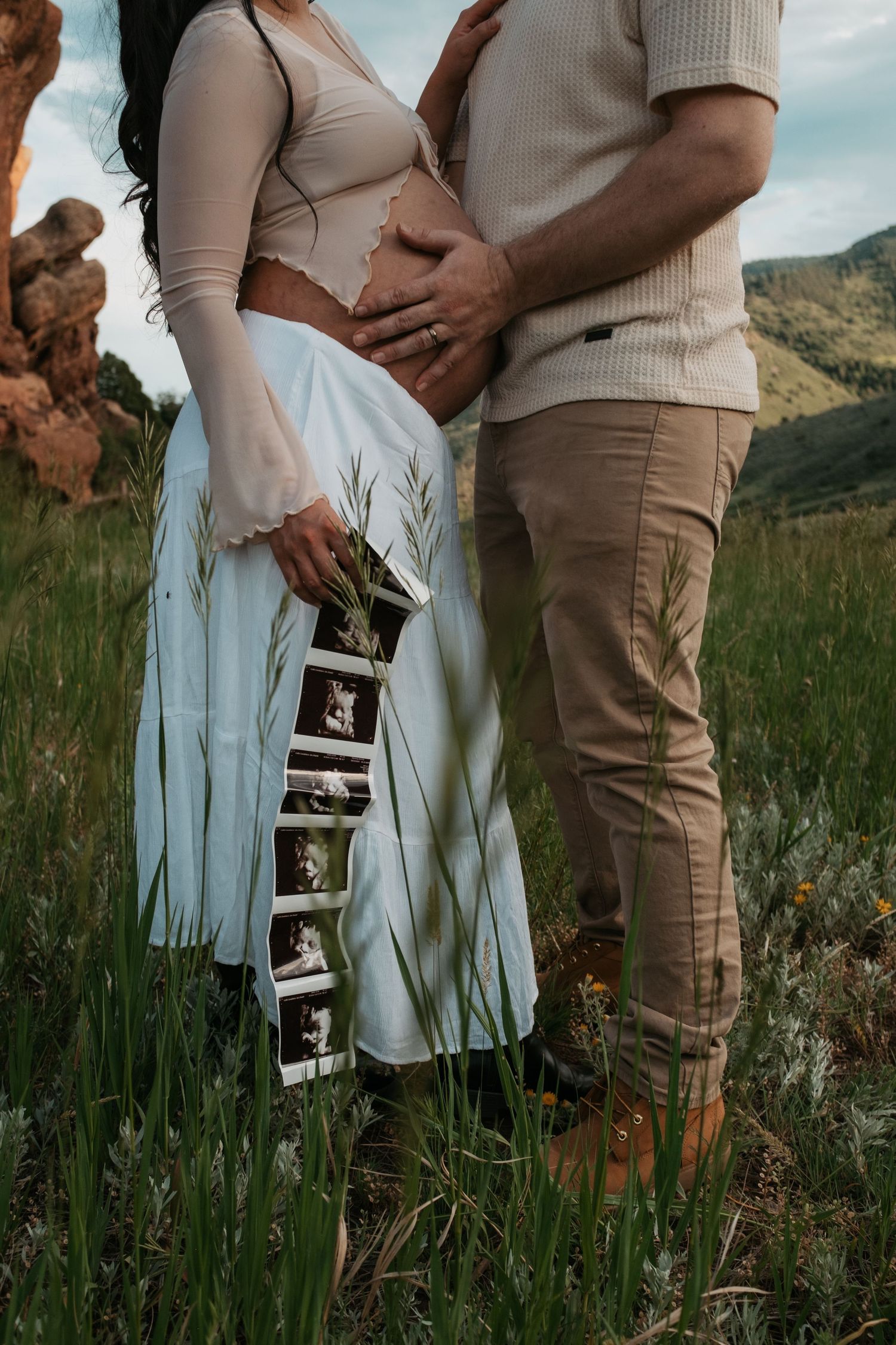 Close-up of a pregnant belly with ultrasound photos hanging against a light blue dress in nature.
