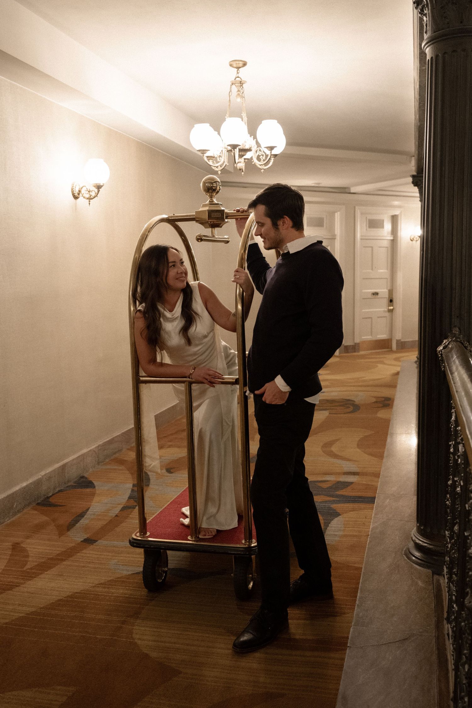 A couple shares a playful moment beside a gold luggage cart in an elegant hotel corridor.