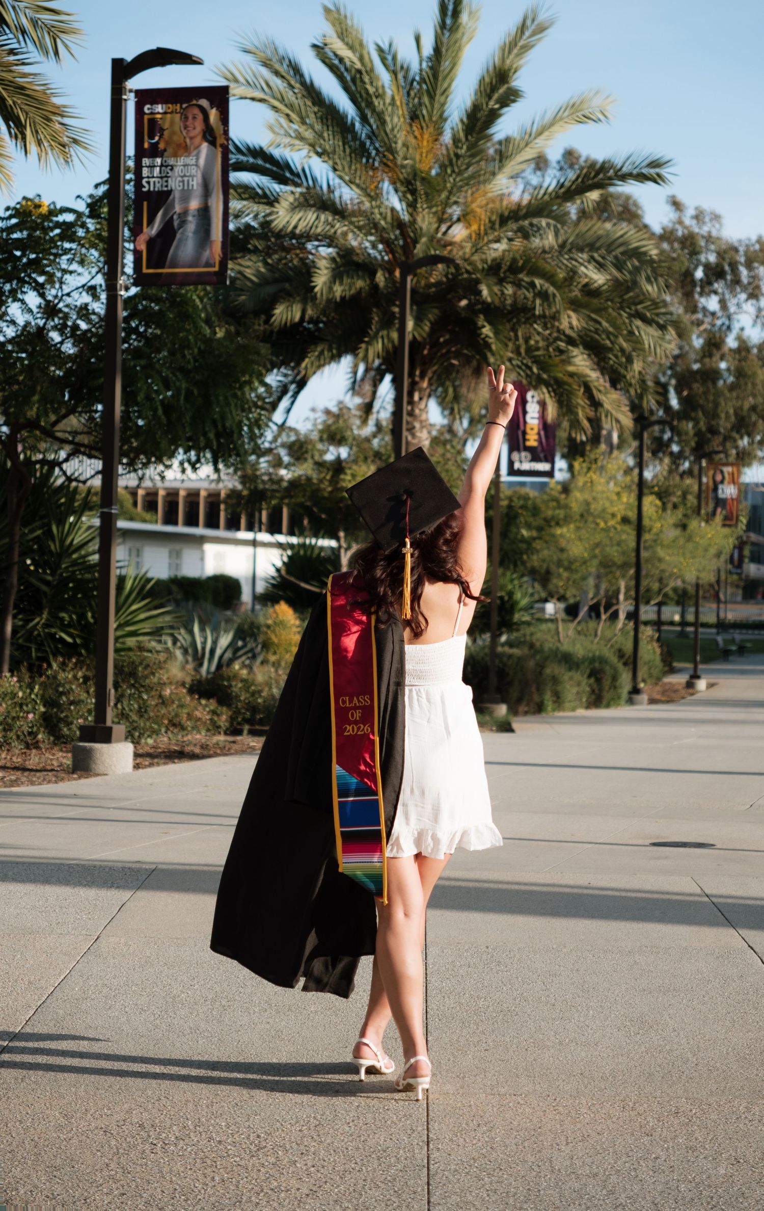 A graduate in white dress holds cap aloft, wearing colorful stole, posing near palm trees on sunny campus walkway.