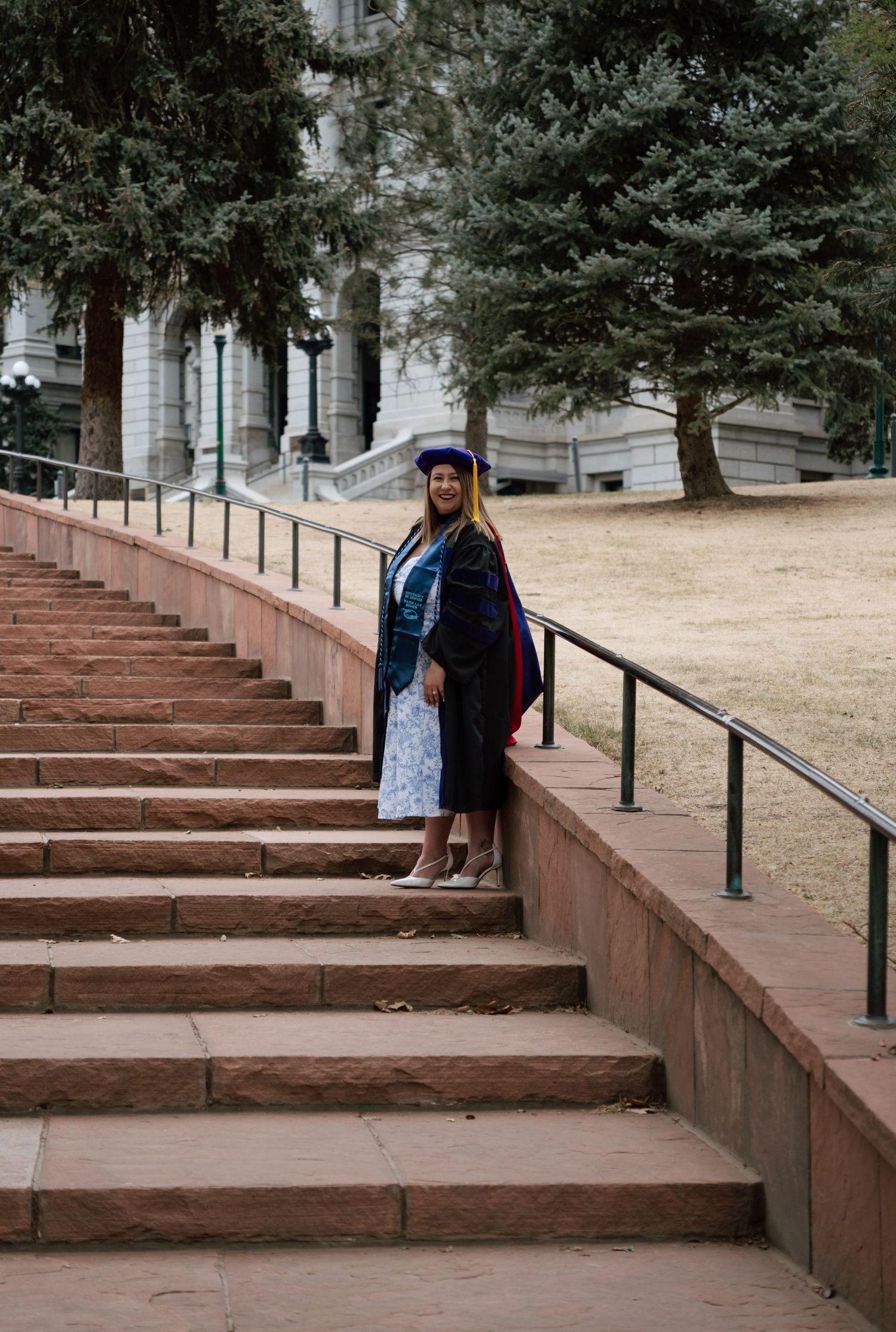 Graduate in cap and gown poses on red brick stairs outside a university building surrounded by evergreen trees.