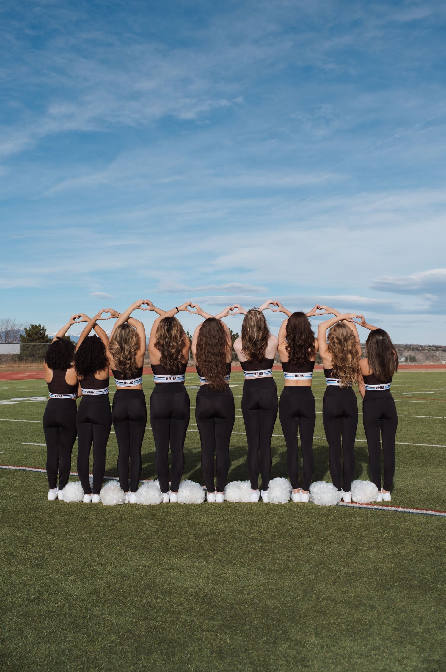 Eight cheerleaders in black uniforms stand on a football field, facing away, forming heart shapes with their hands.