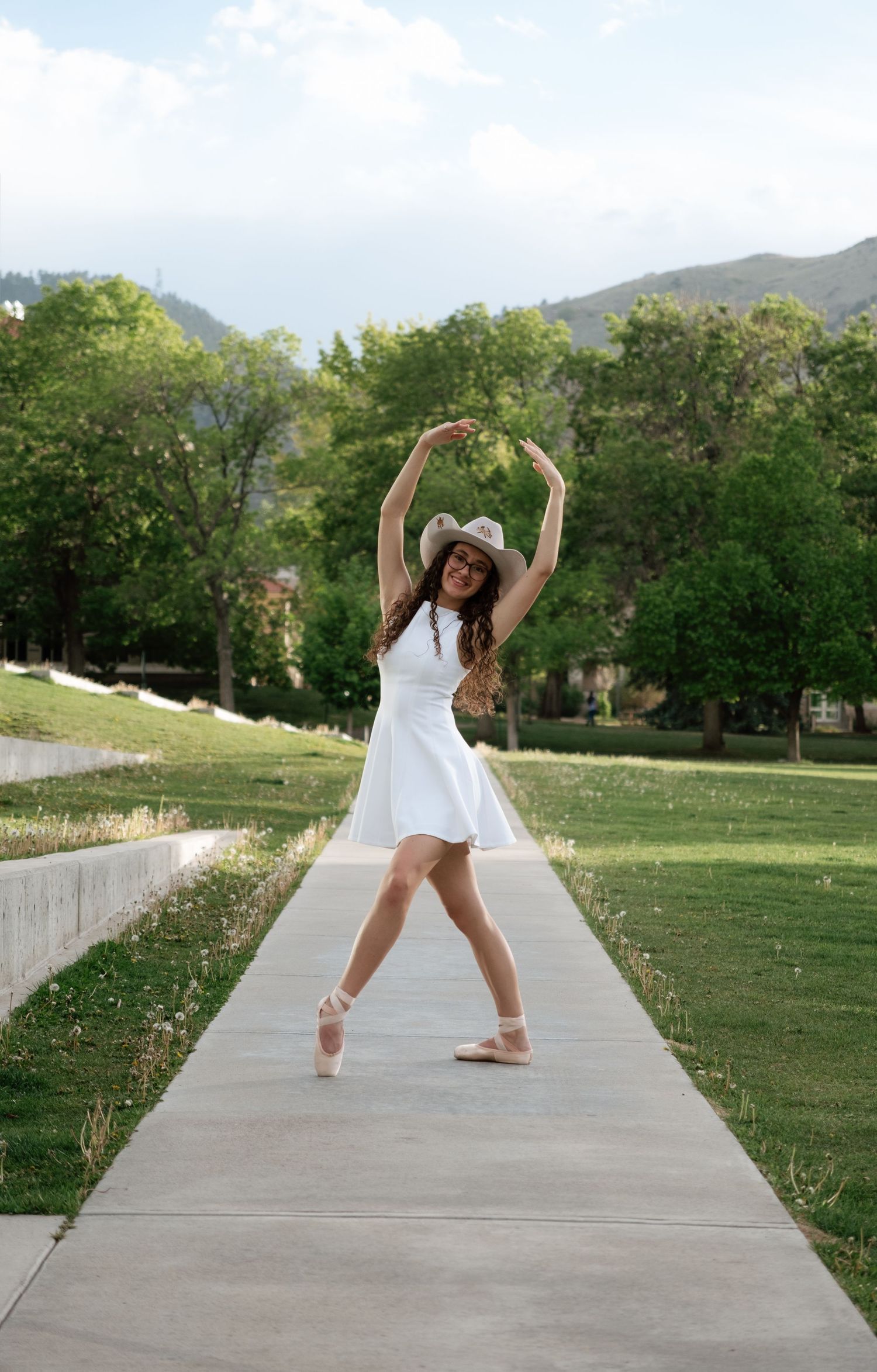 Ballerina in white dress and cowboy hat poses on tiptoe on a park path with mountains and trees behind.