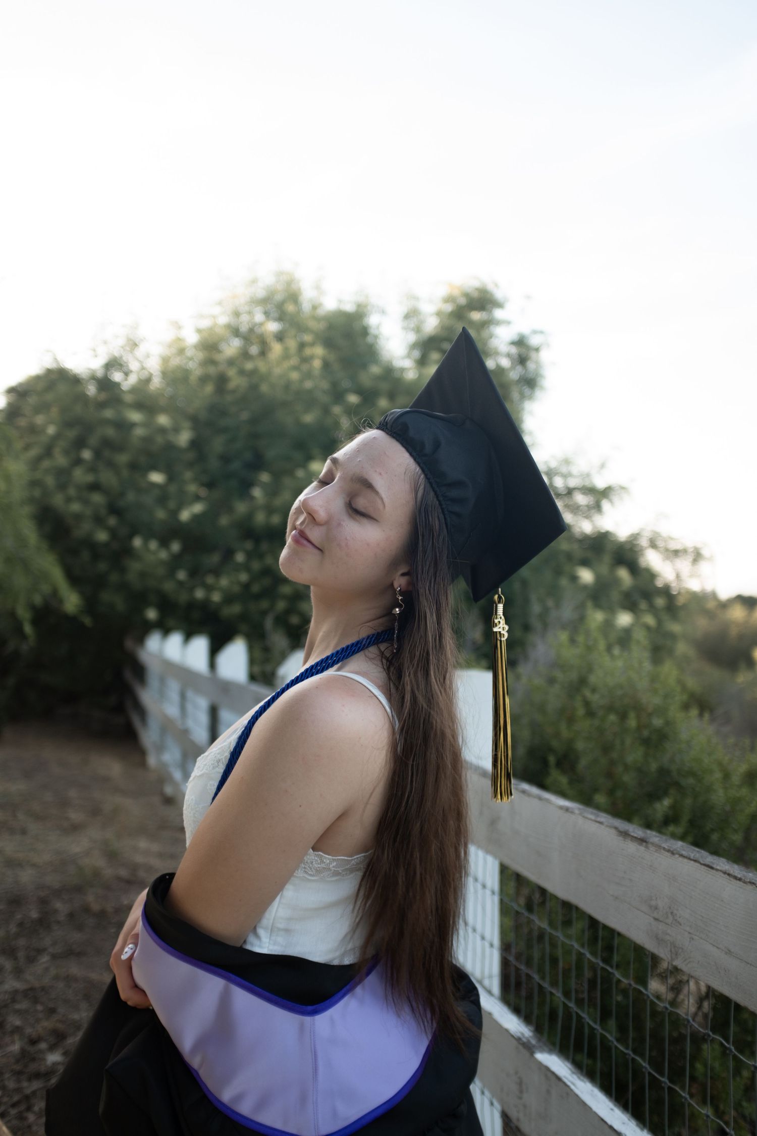 Graduate in academic regalia poses against white fence during golden hour.