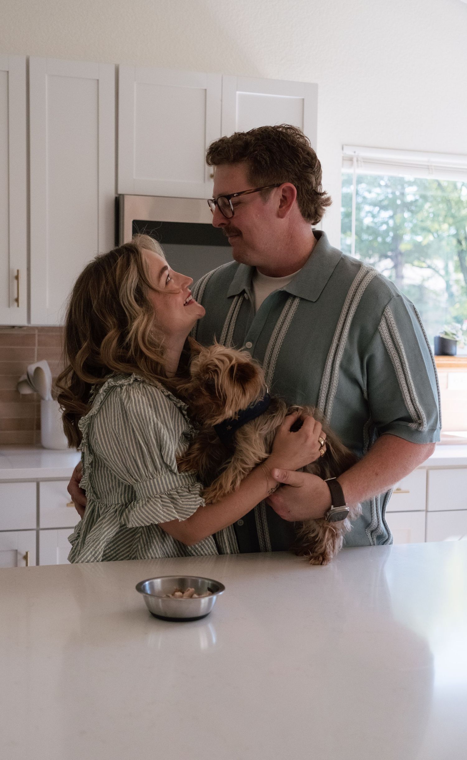 Smiling couple embracing in a bright modern kitchen while holding their small Yorkshire terrier, with a dog bowl on the white countertop.