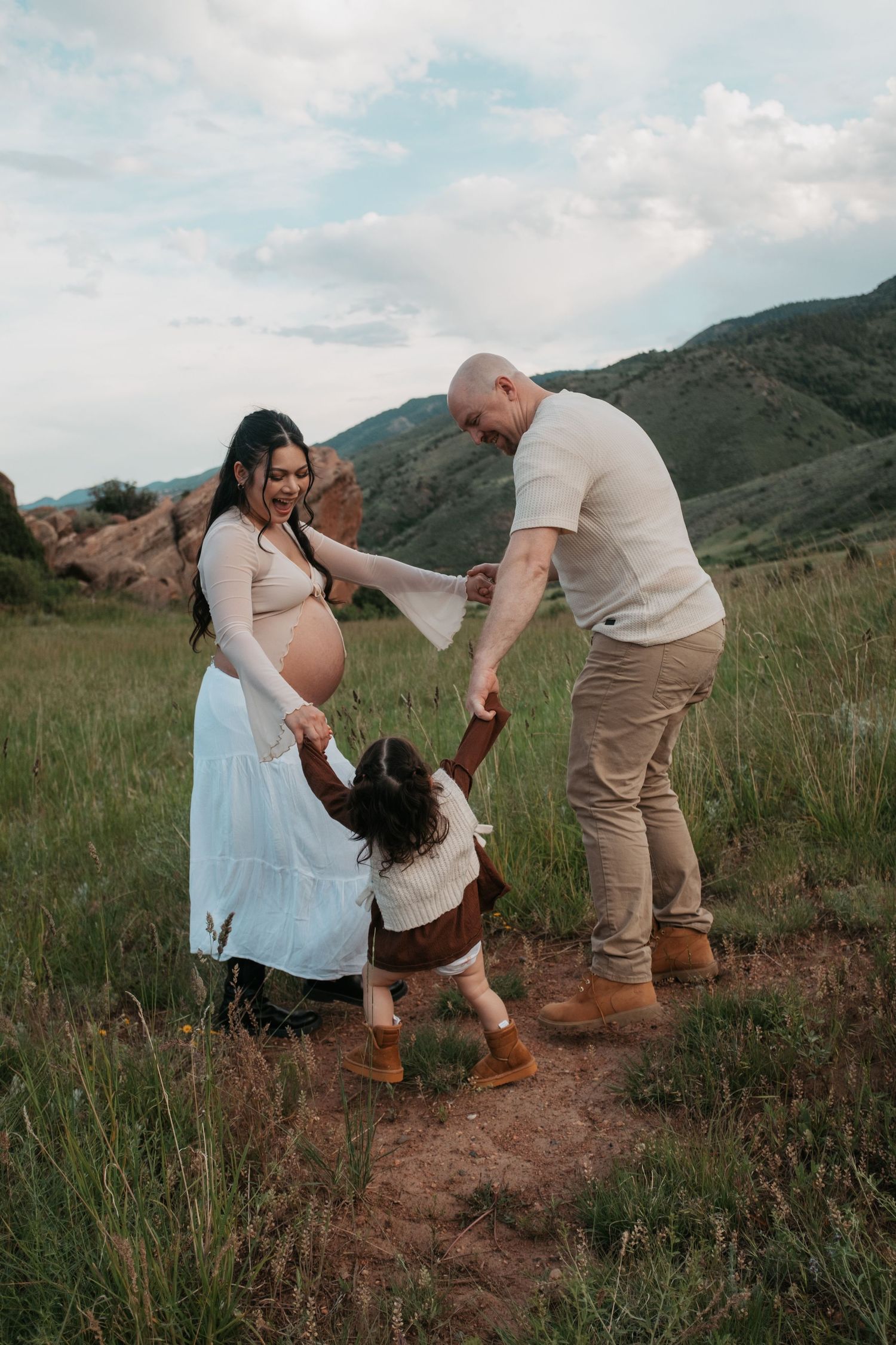 Family celebrating pregnancy with joyful dance moves in a mountain meadow.