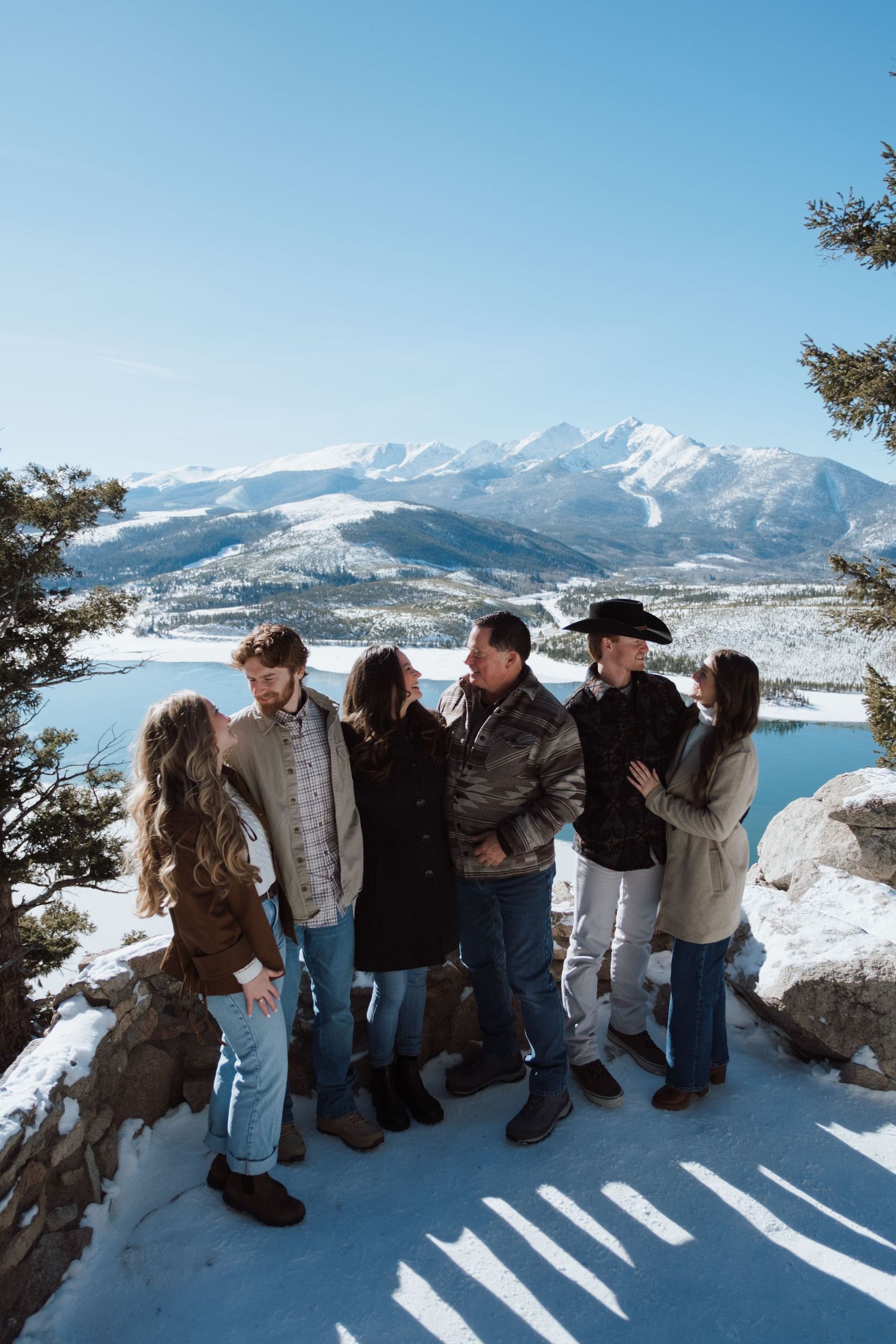 Group of six friends posing on snowy overlook with mountain lake and blue sky backdrop in winter.