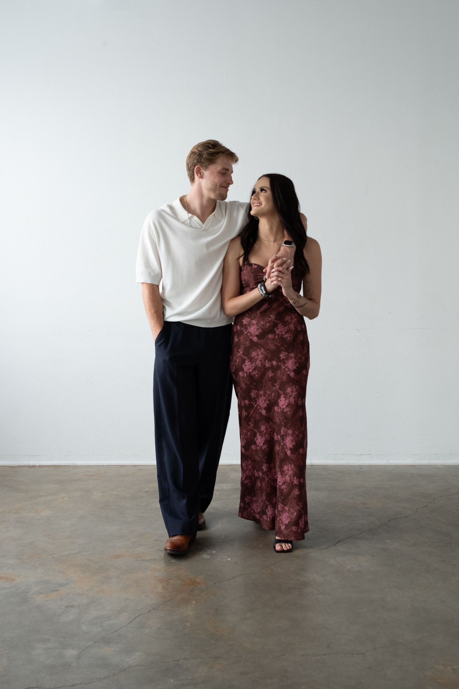 A couple in elegant attire smiling at each other, man in white shirt and black pants, woman in floral maxi dress.