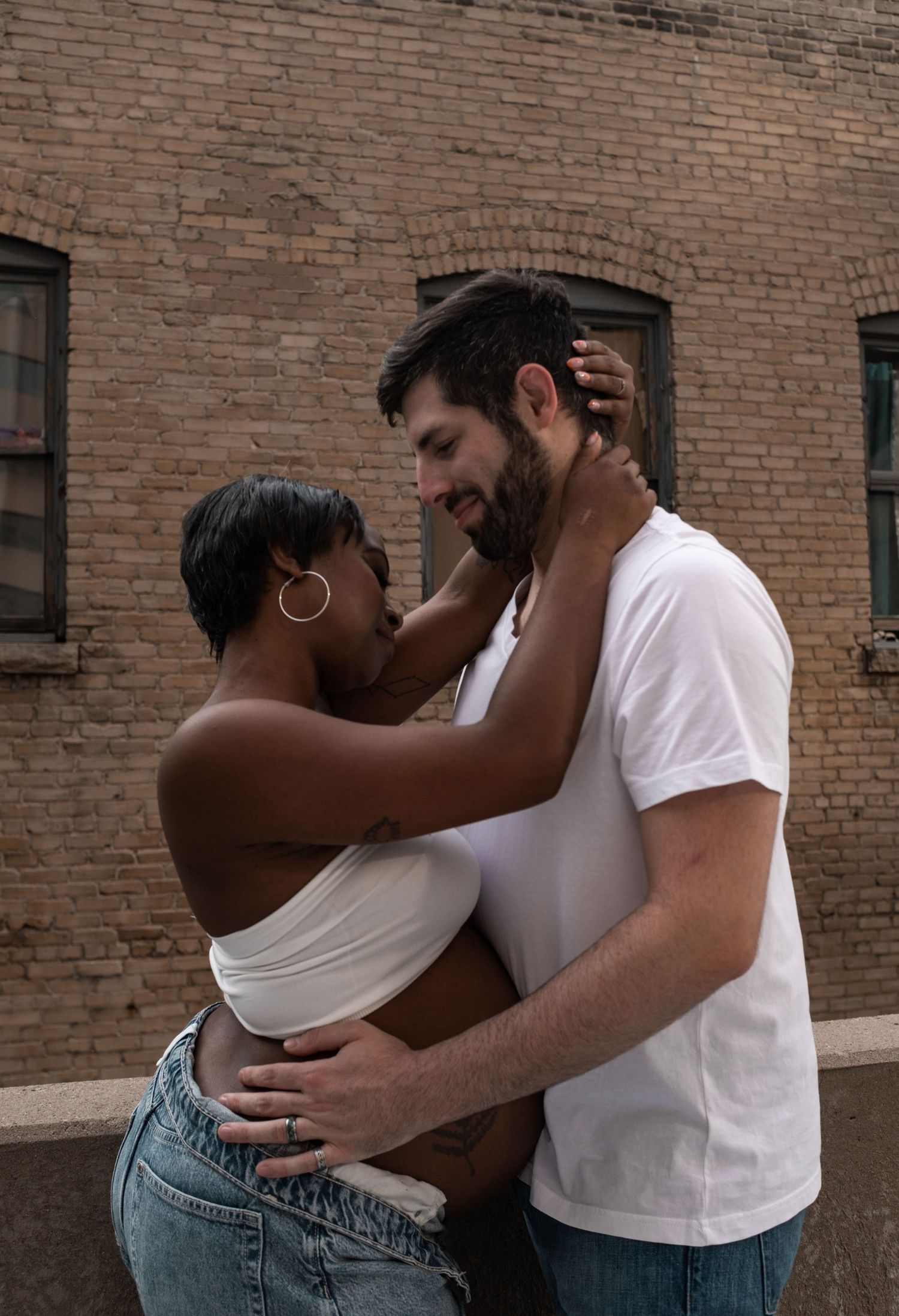 An interracial couple embraces romantically in front of a brick wall, sharing a tender moment outdoors.