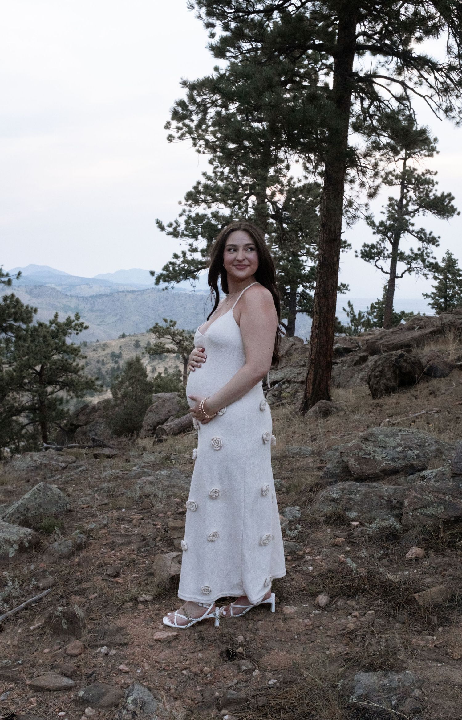 Pregnant woman in white floral dress cradling baby bump on rocky mountain hillside with pine trees.