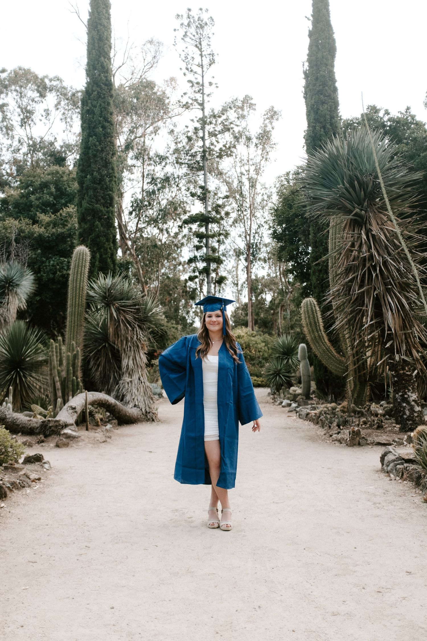 Graduate in blue academic regalia poses on path surrounded by desert plants.