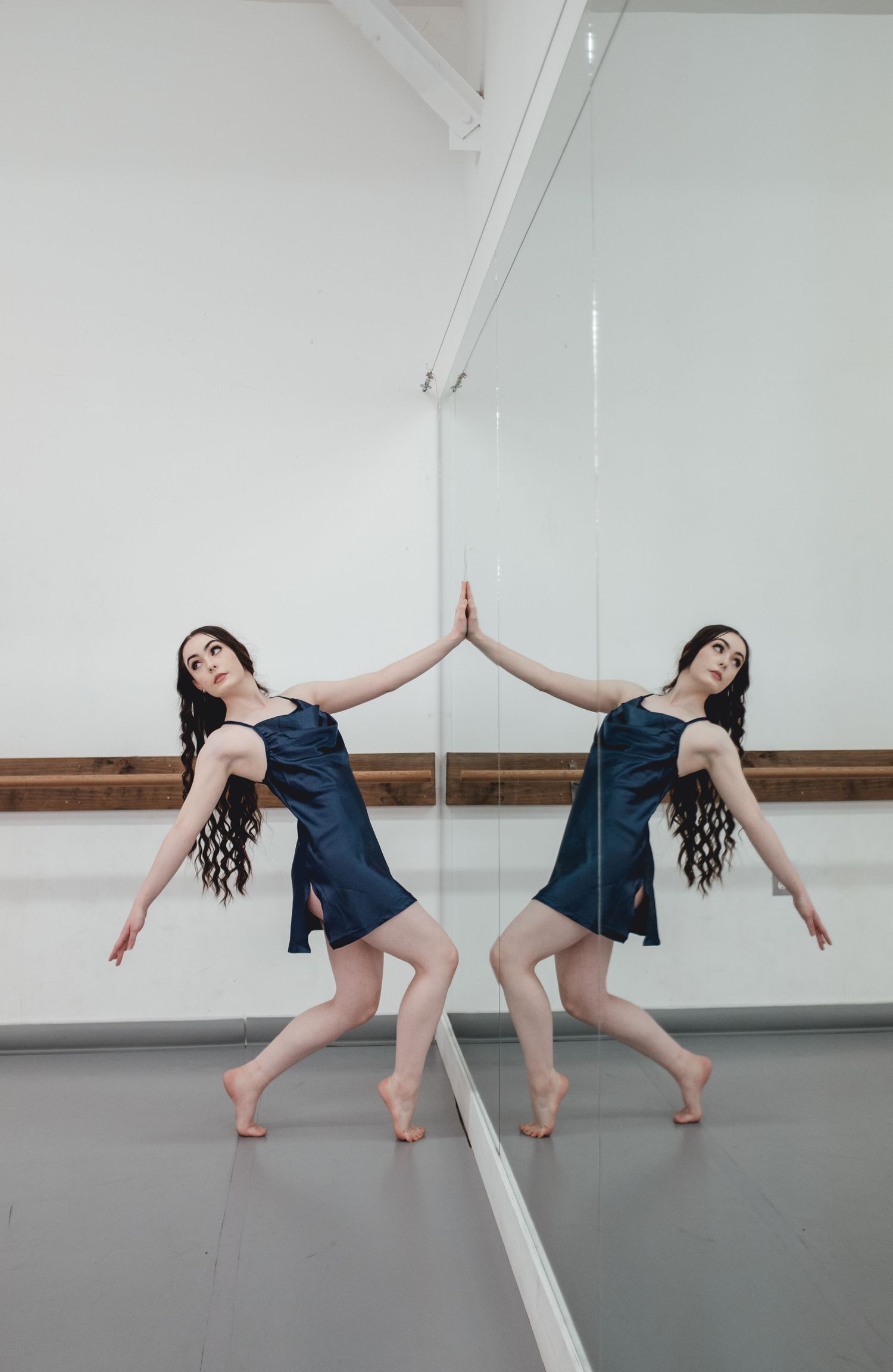 Dancer in navy dress performs dynamic movements in white studio with mirror.