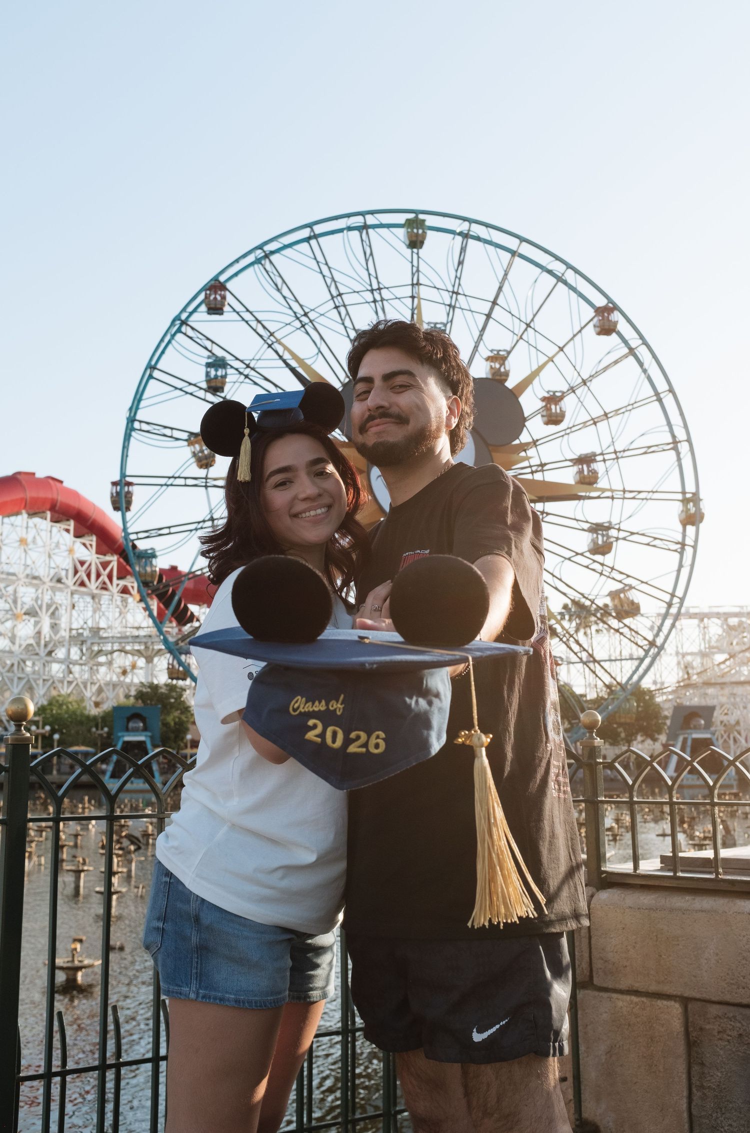 Couple posing with Mickey Mouse graduation hats at Disneyland California Adventure with Ferris wheel backdrop.