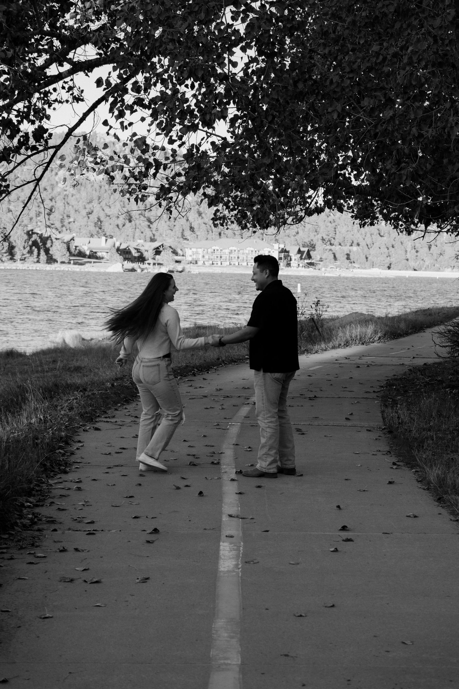 Black and white photo of a couple holding hands on a lakeside path under a tree.