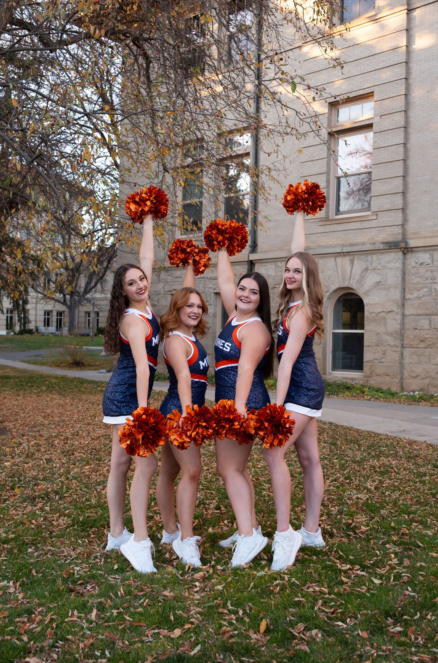 Four cheerleaders in navy and orange uniforms pose with pom-poms outside a stone building on an autumn day.