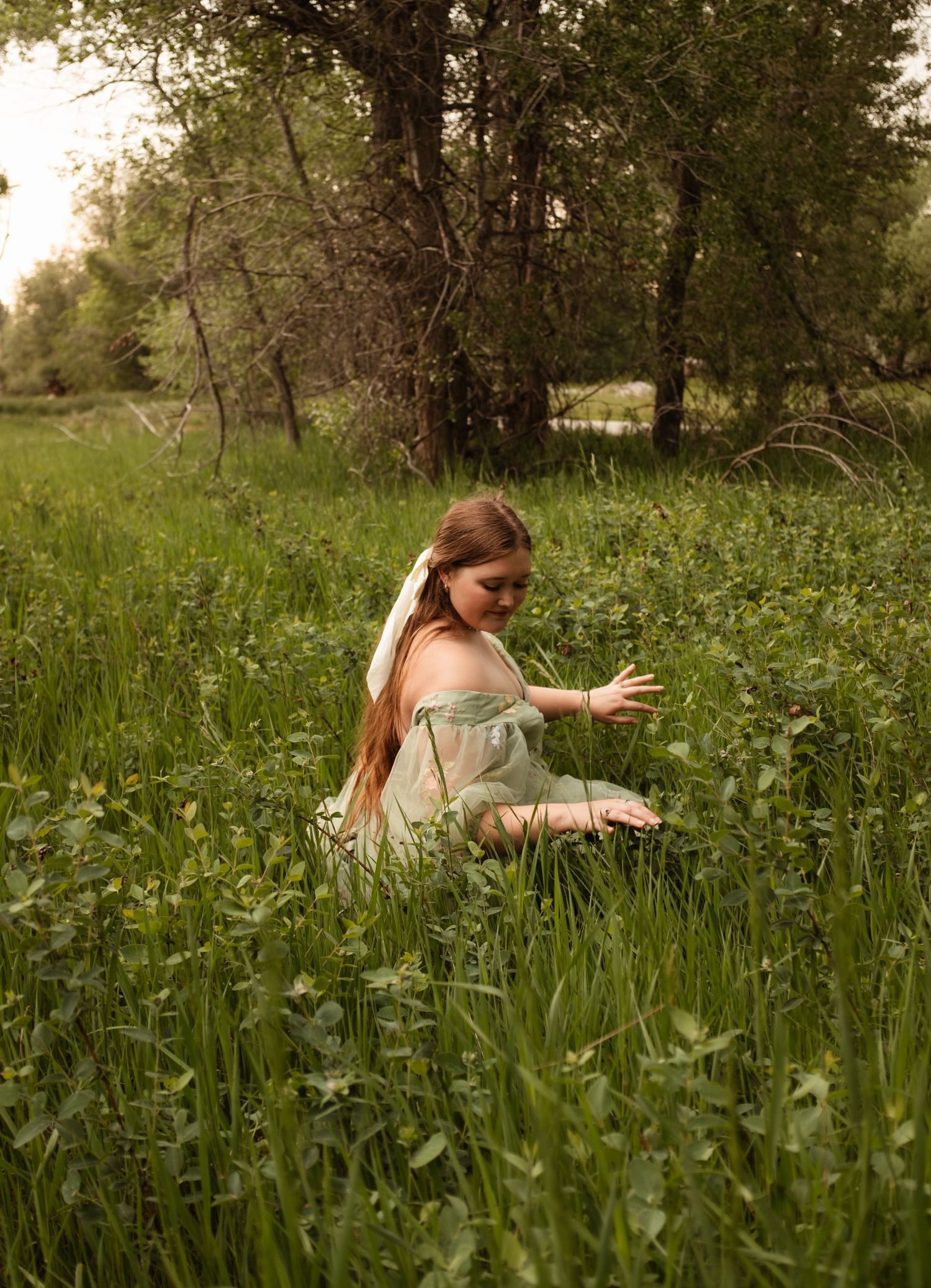 Figure in an off-shoulder dress reaching for wildflowers in a lush summer meadow.