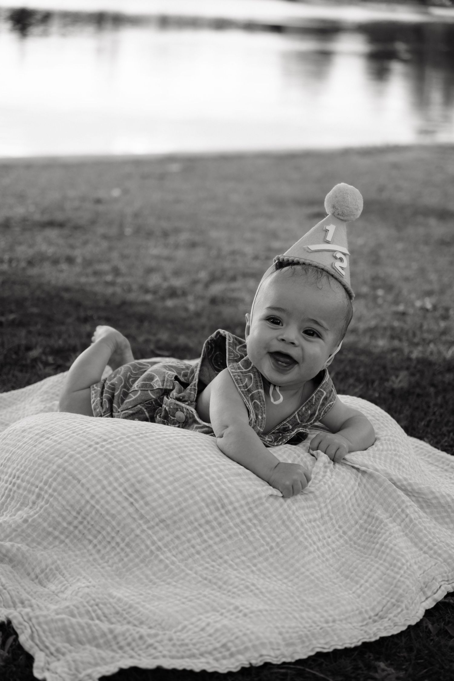 Black and white photo of a smiling baby wearing a birthday hat, lying on a blanket near a lake.