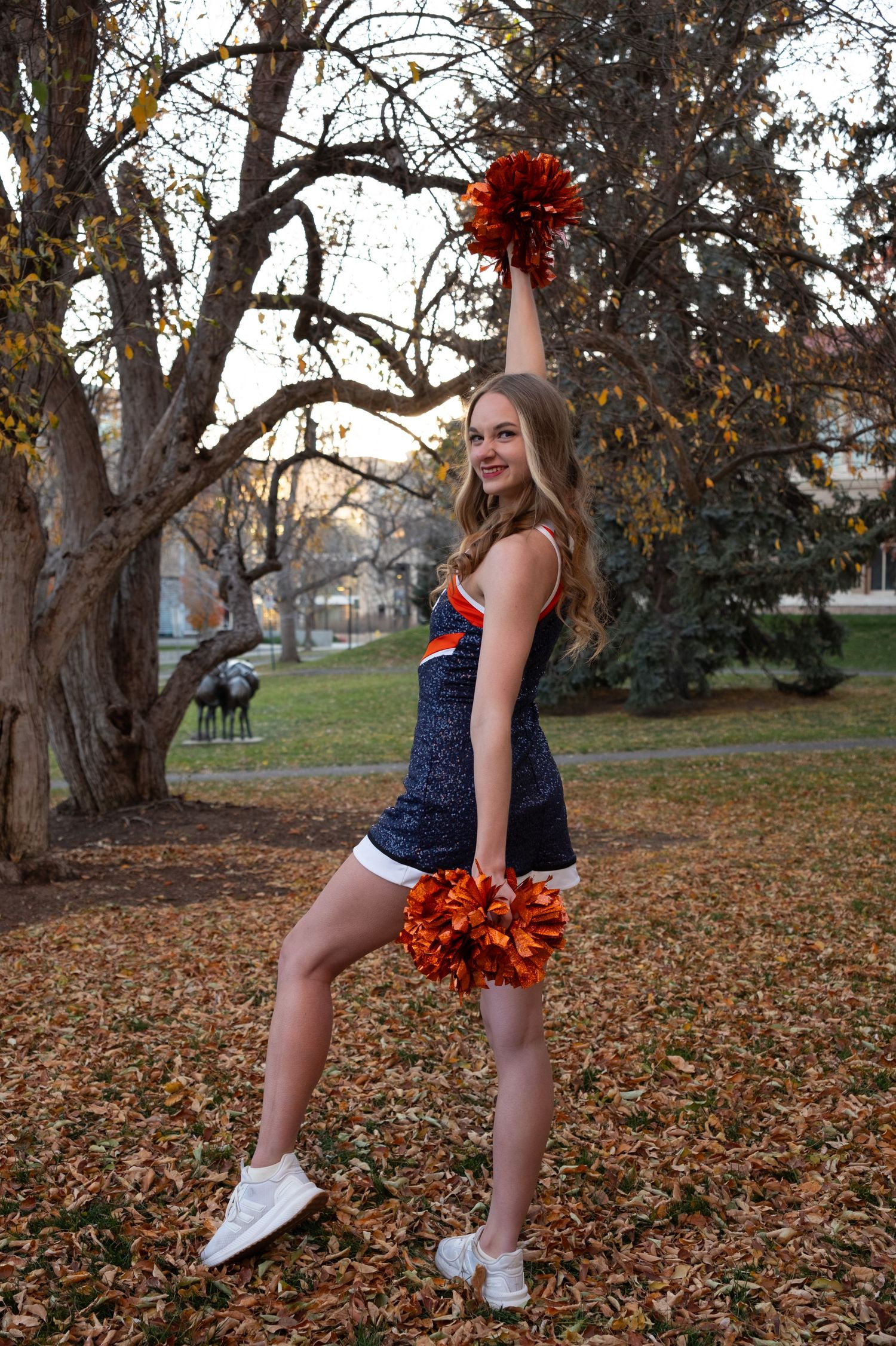 Cheerleader in navy and orange uniform poses with pom-poms outdoors among autumn trees and fallen leaves.