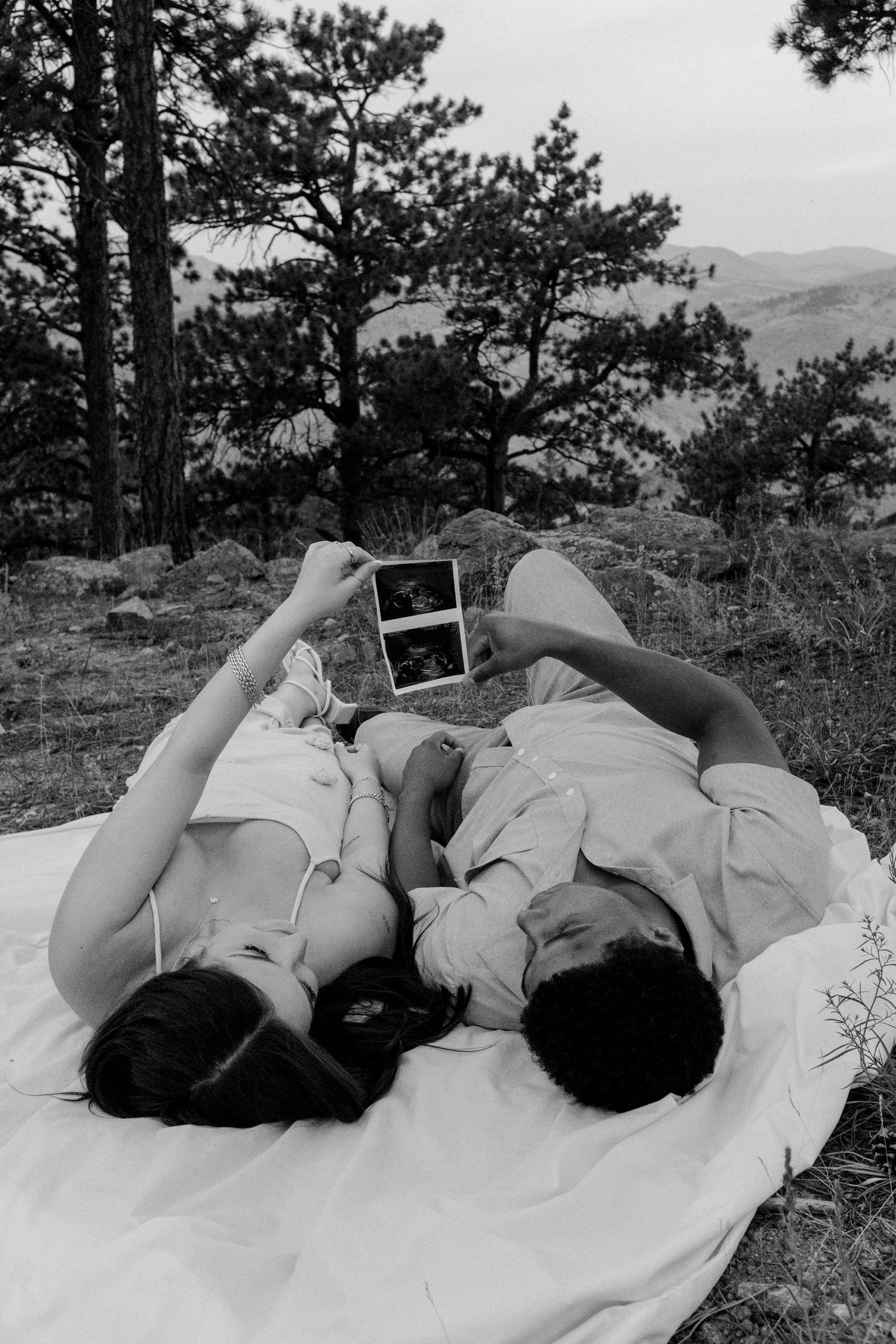 Black and white photo of a couple lying on a blanket outdoors, holding up an ultrasound photo together.