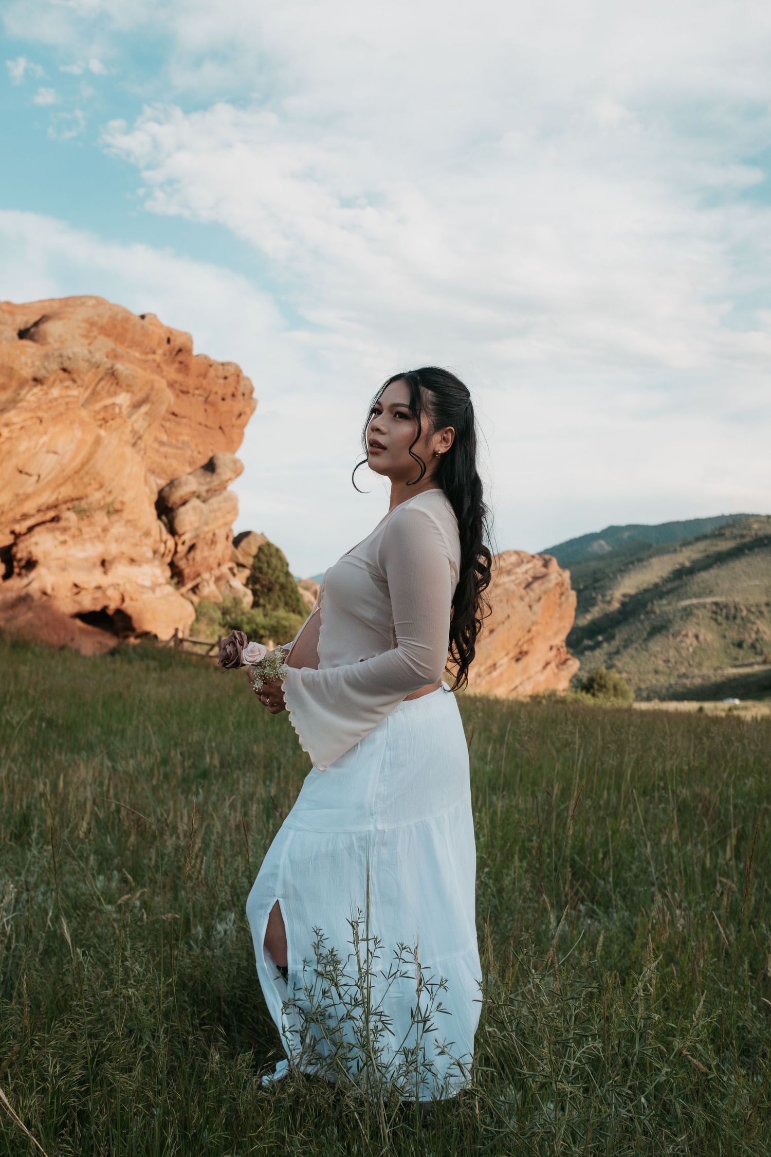 Maternity photo shoot in a flowing white dress against red rock formations and mountain landscape.