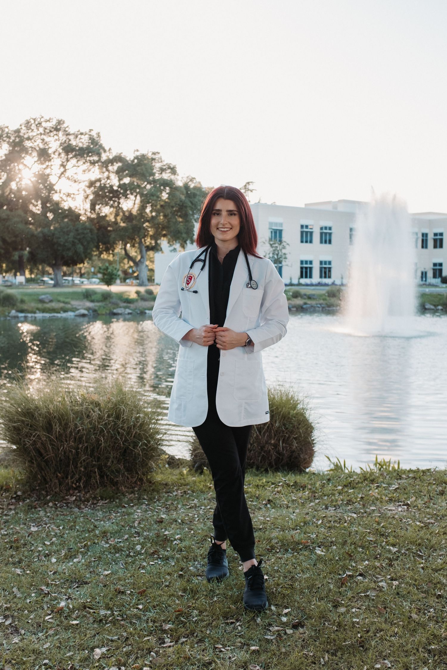 Graduate in white coat standing by a fountain on campus grounds.