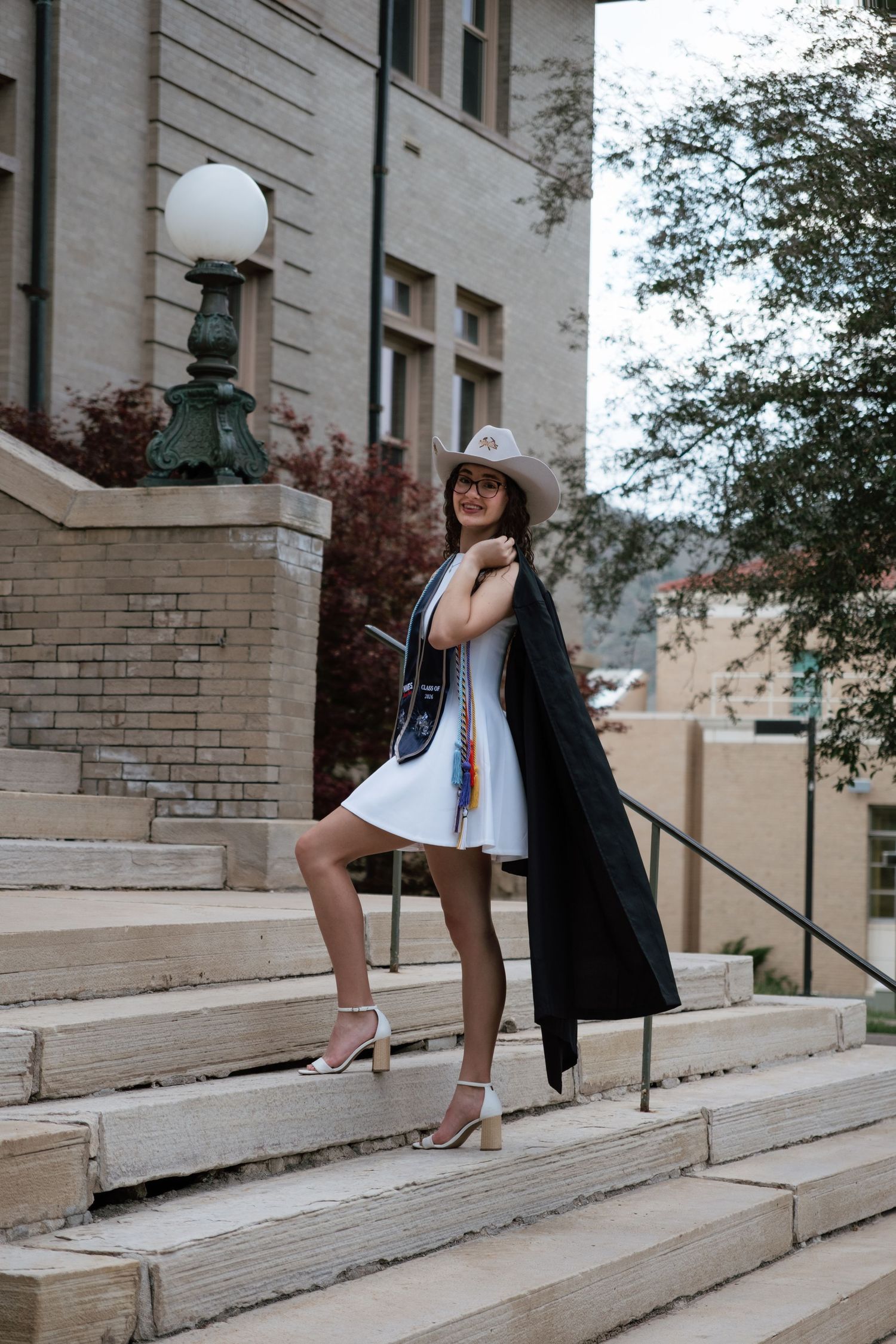 Woman in white dress, graduation gown, and hat poses on university steps in heels.