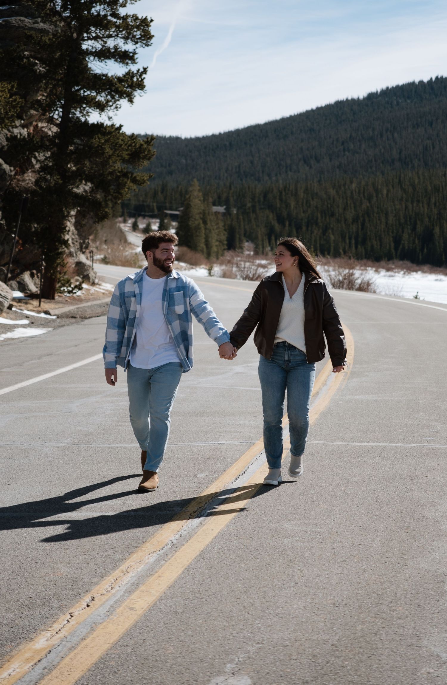 Couple holding hands walking on a mountain road surrounded by pine trees and snow in casual winter outfits.
